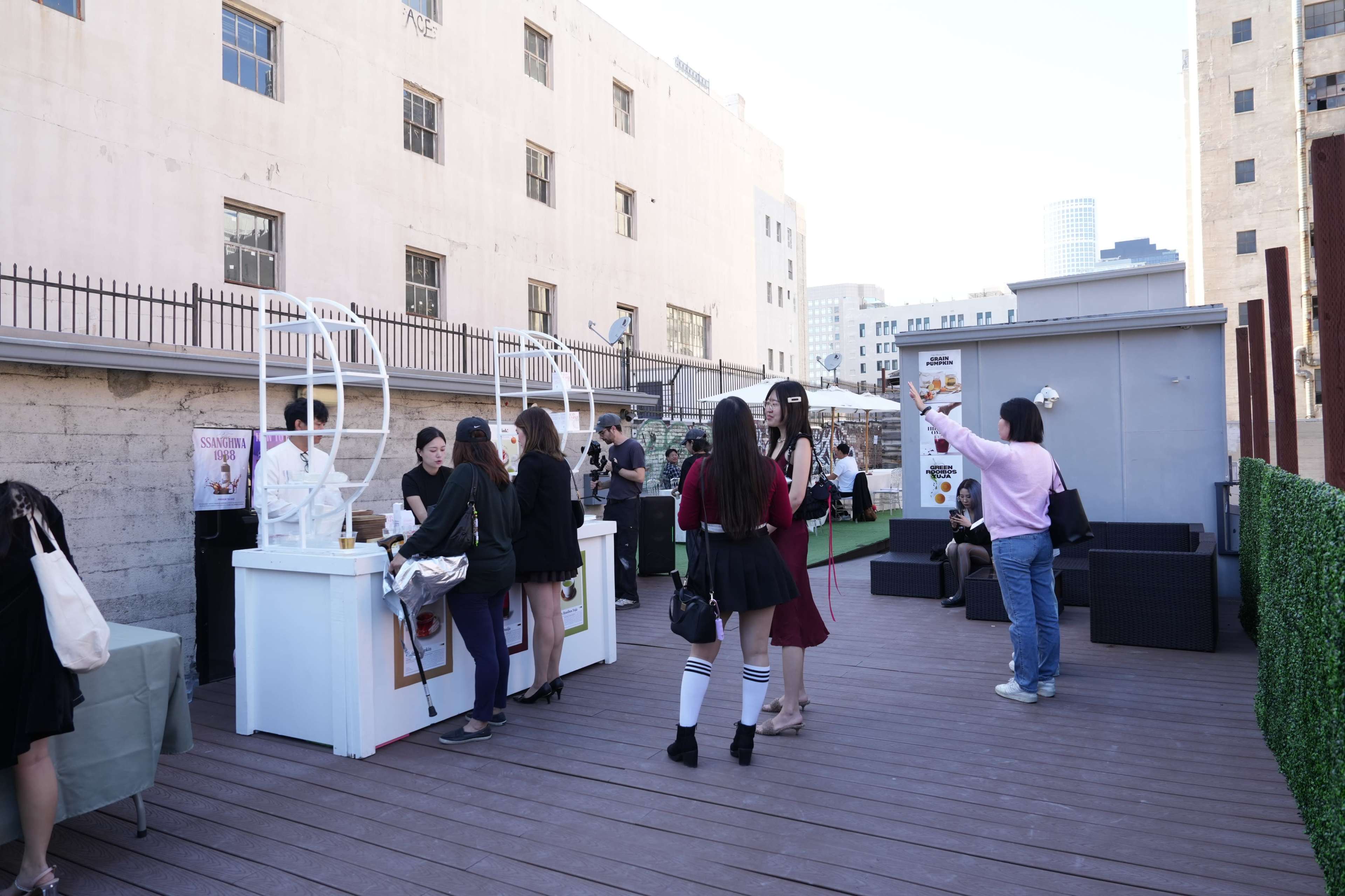 A rooftop scene features several people socializing, with a few kiosks serving food or drinks and a backdrop of urban buildings.