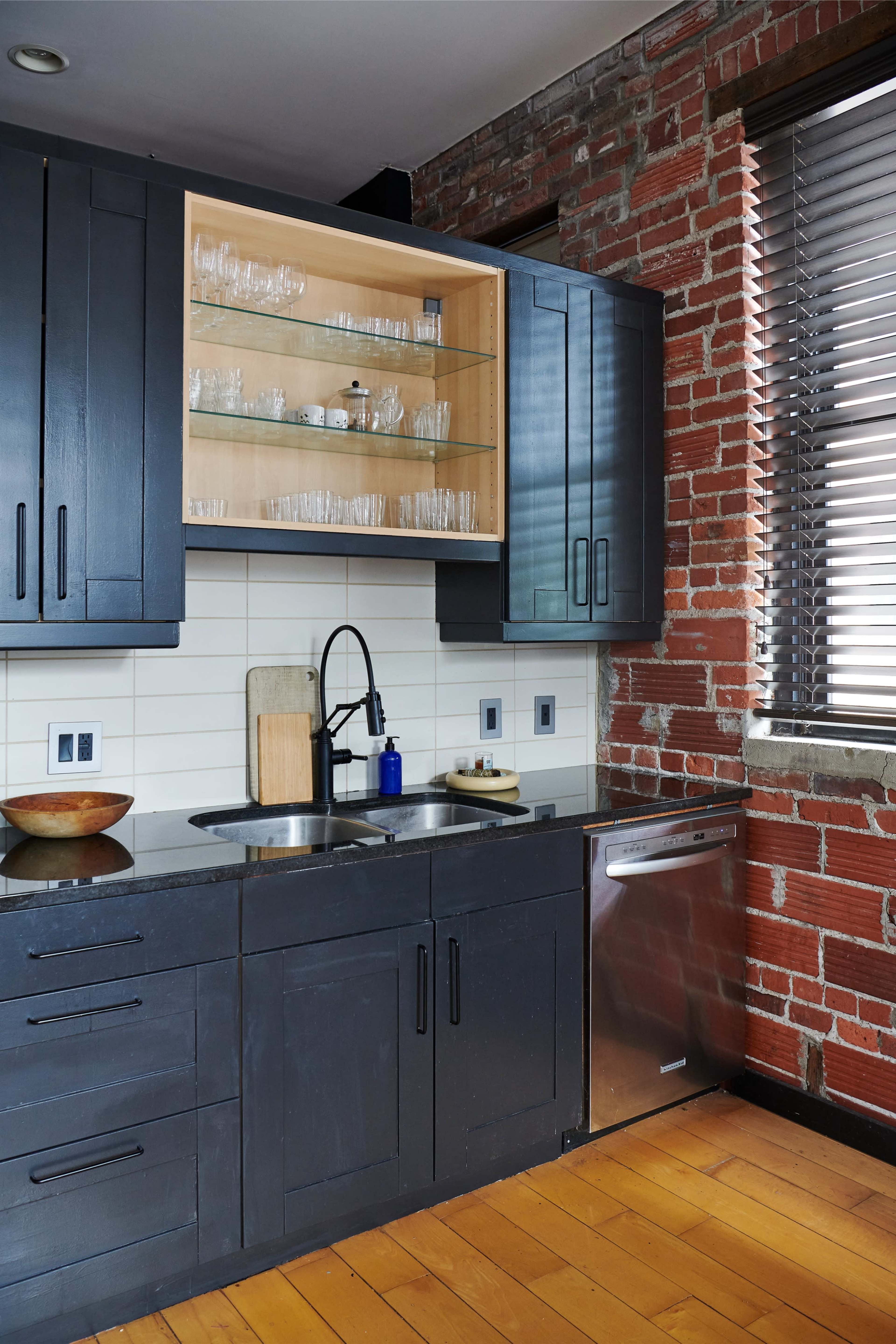 A modern kitchen featuring dark cabinets, a stainless steel dishwasher, a double sink with a black faucet, and an exposed brick wall.