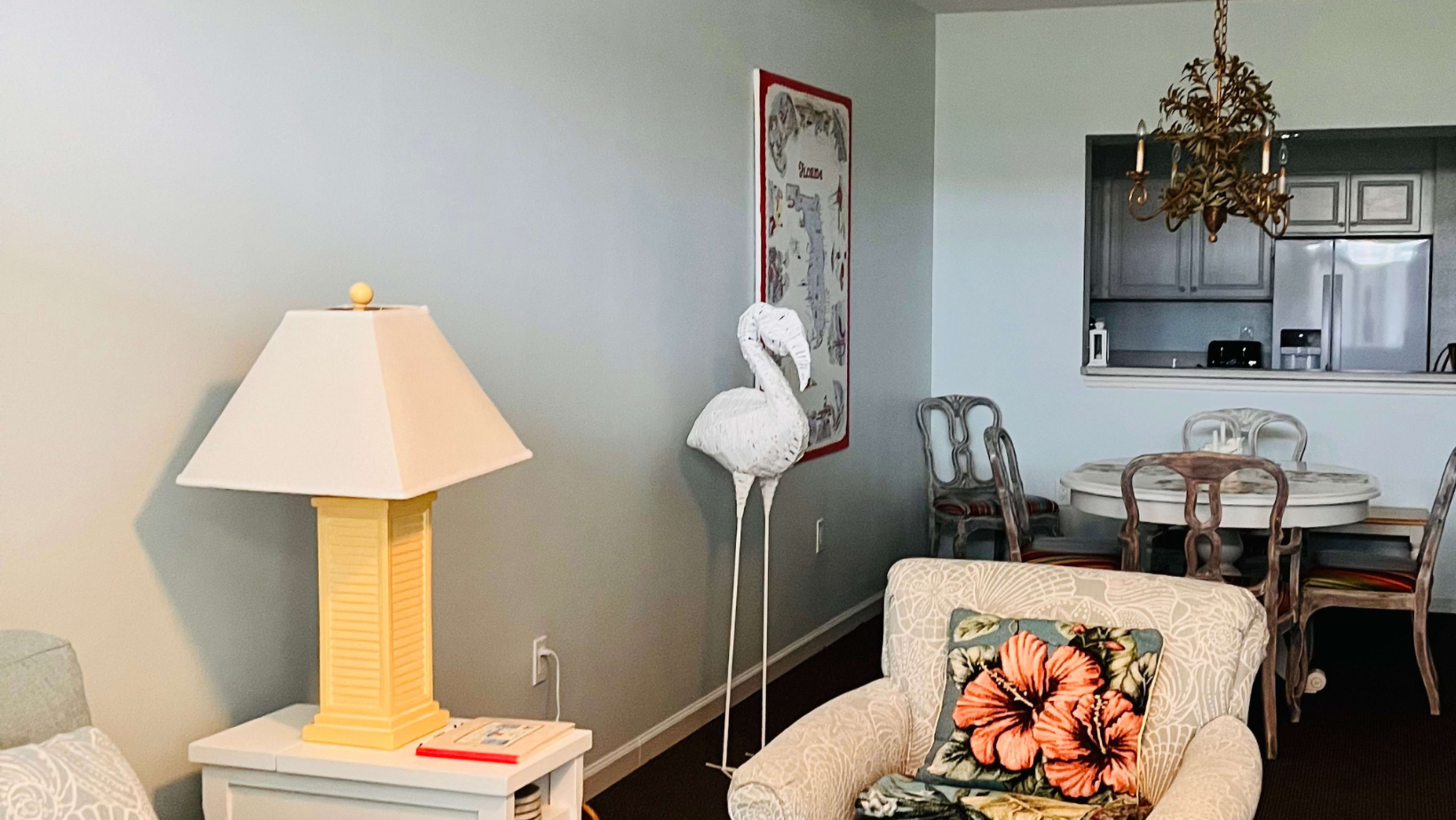 A light-colored living room featuring a patterned armchair, a lamp, a flamingo decoration, and a dining area in the background with a round table and chairs.
