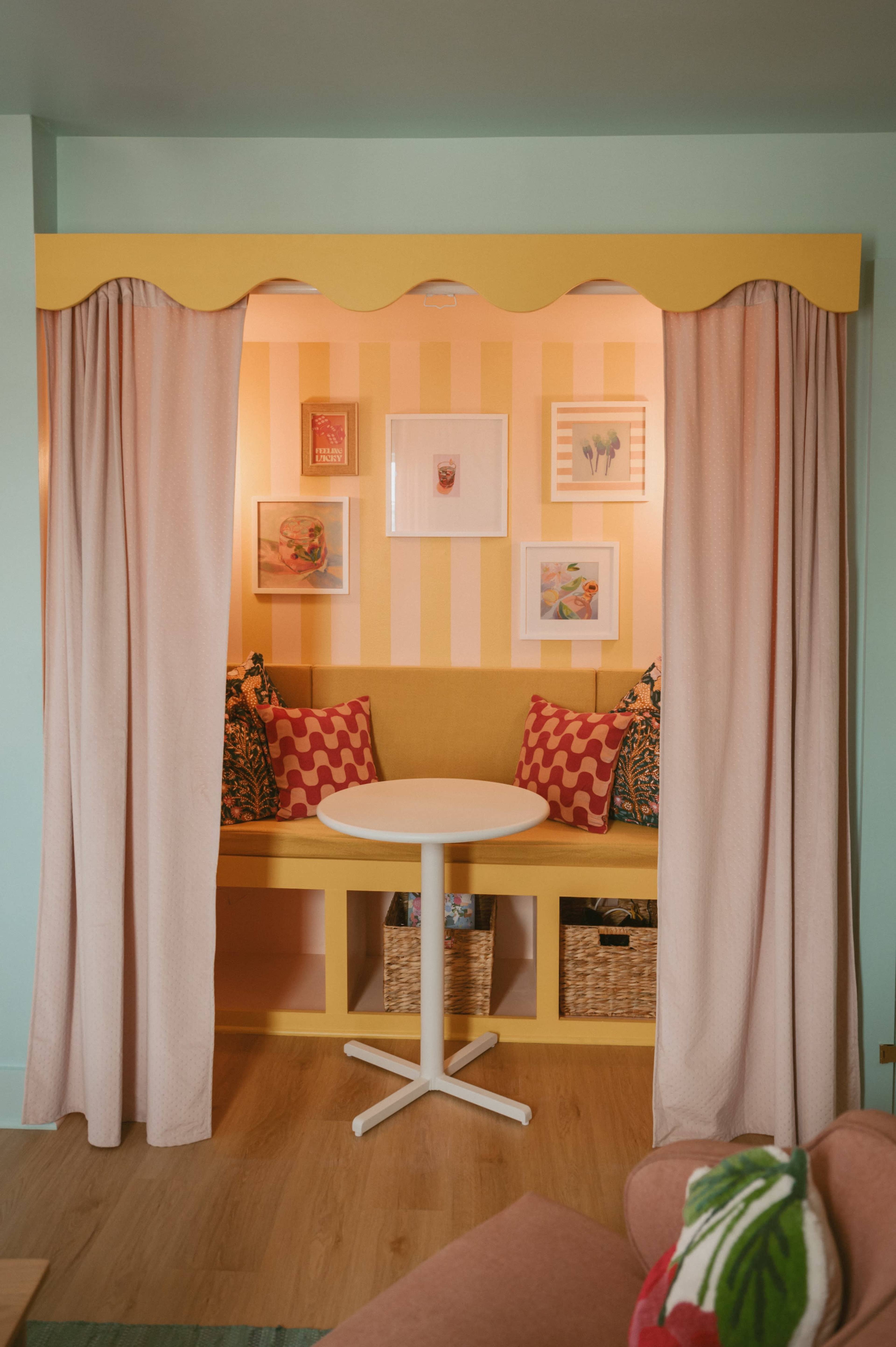A cozy nook with a yellow and white striped wall, featuring a round table, built-in seating with patterned cushions, and framed artworks on display.