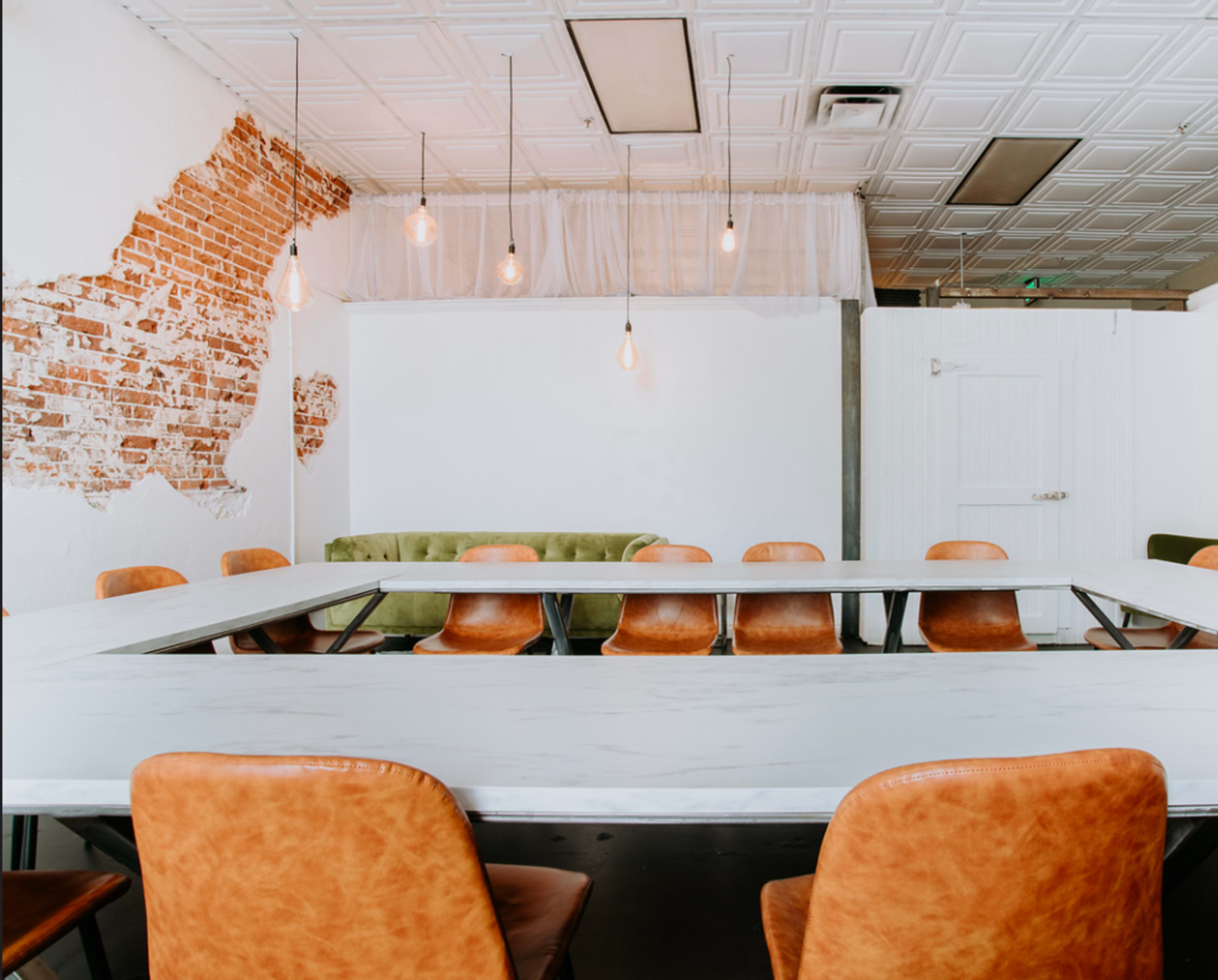 The image shows a large meeting room with a long table surrounded by leather chairs, a green couch against a wall, and exposed brick features.