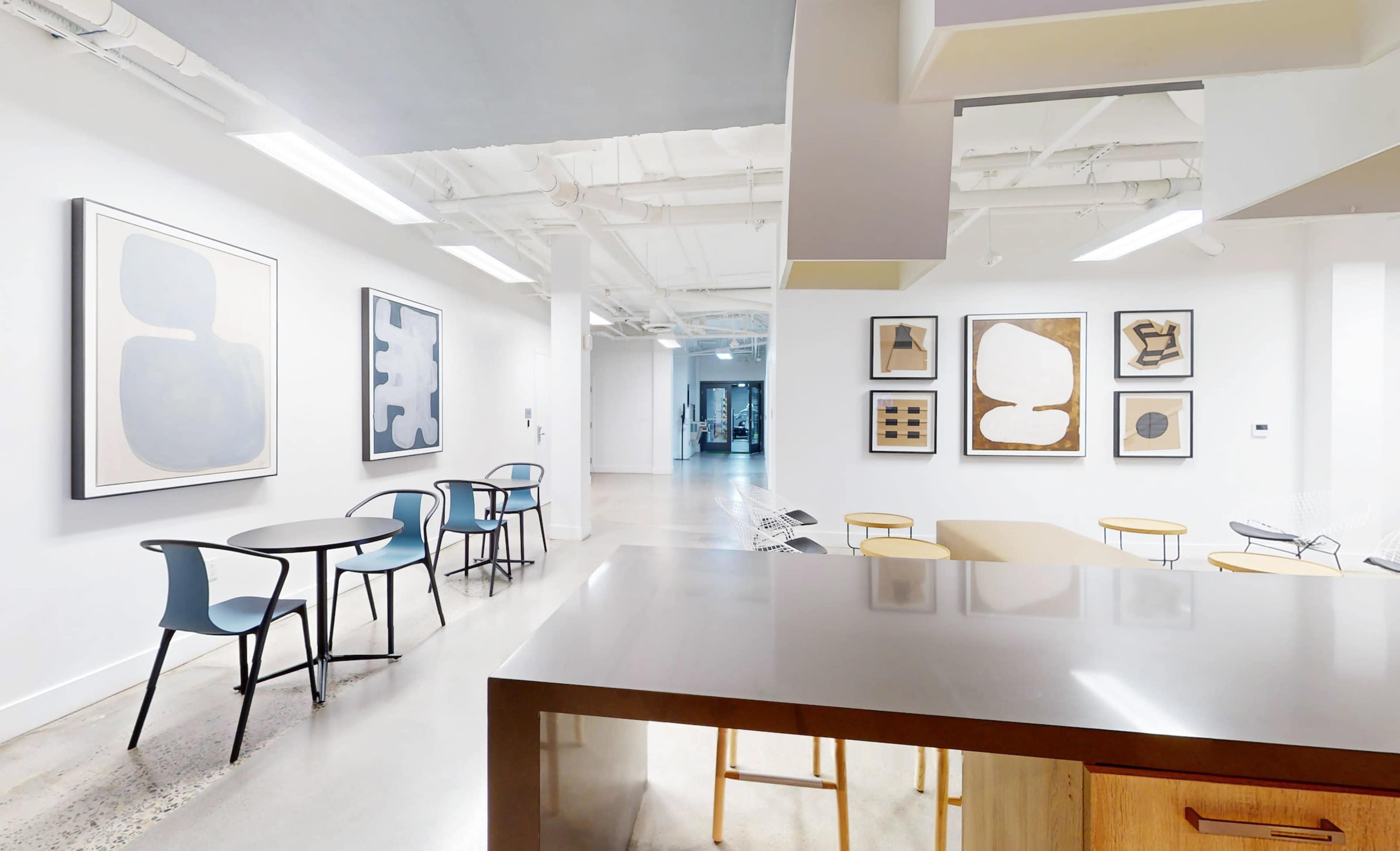 A modern workspace features a kitchen area with a countertop and wooden cabinetry, adjacent to a seating area with black and blue chairs, and walls adorned with framed abstract artwork.