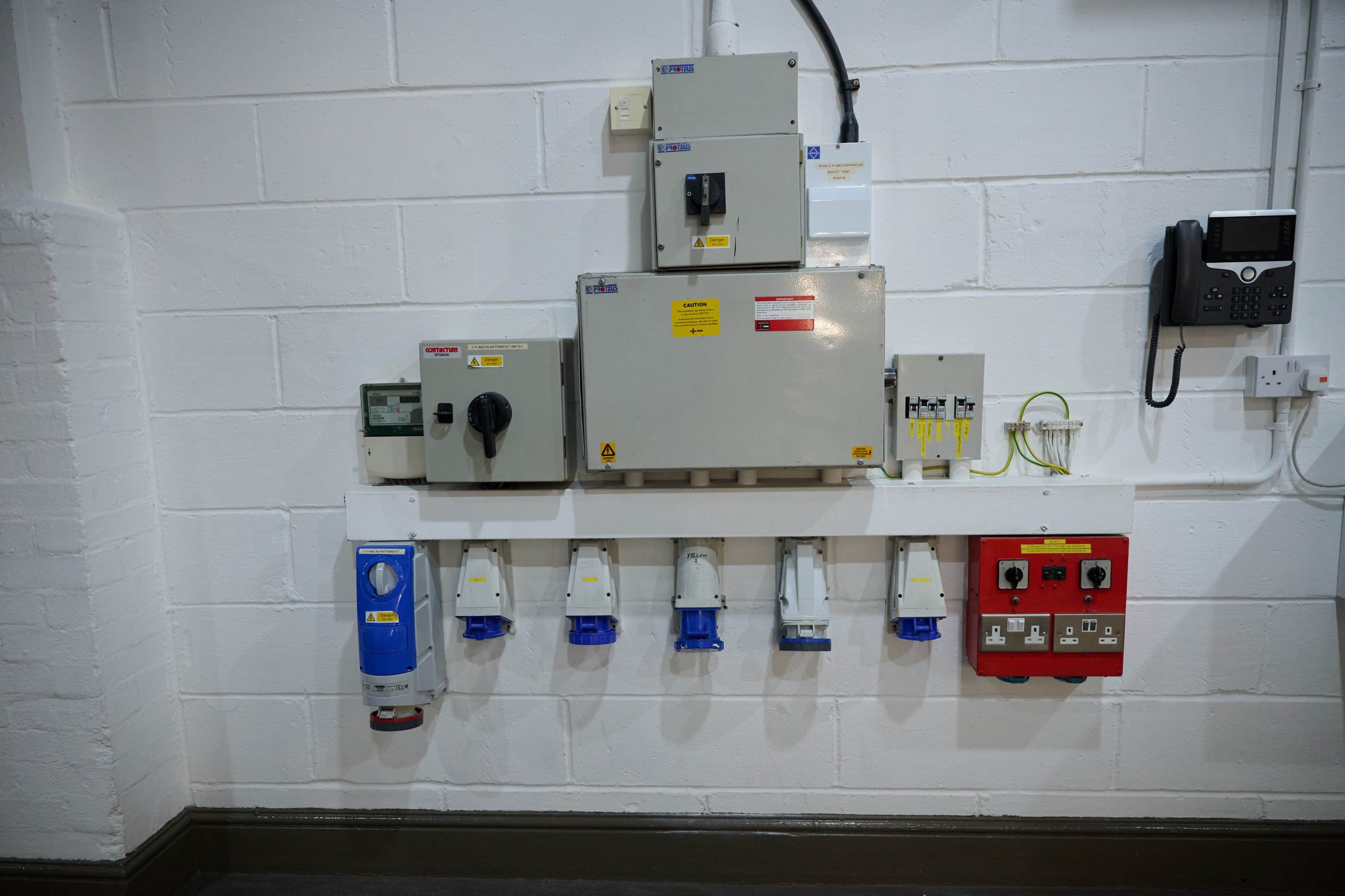 The image shows an industrial electrical panel mounted on a white wall, featuring various circuit breakers, switches, and power outlets.