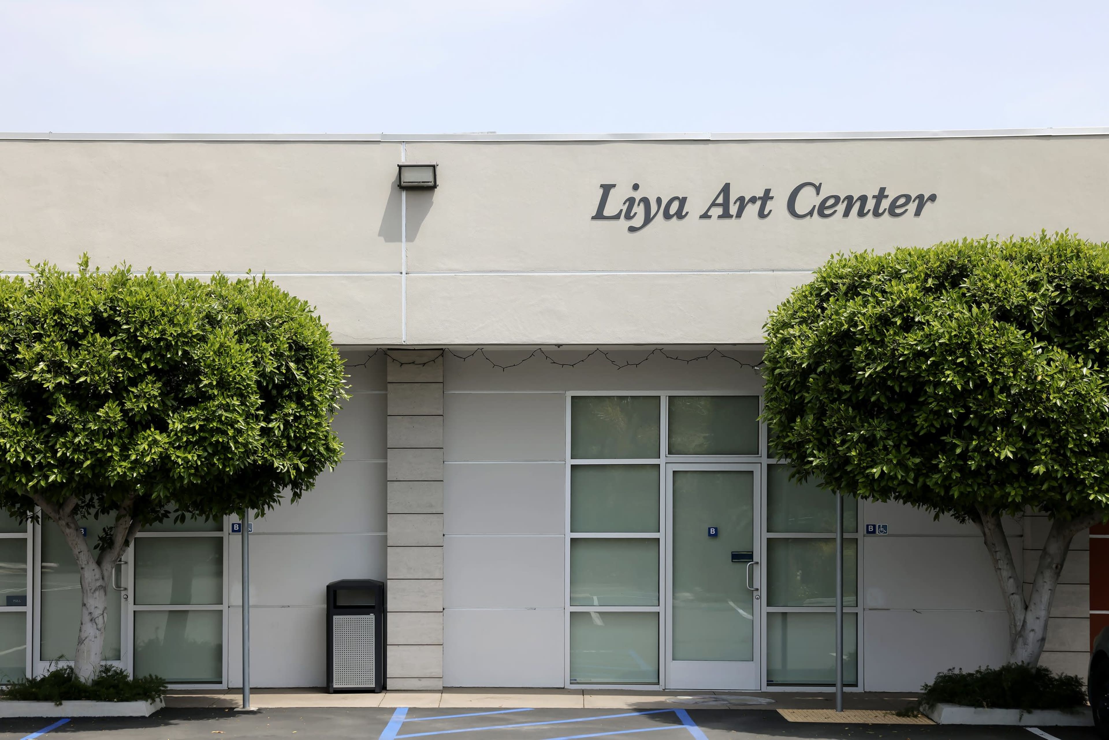The exterior of the Liya Art Center building with two large, rounded shrubs in front and a trash bin to the left.