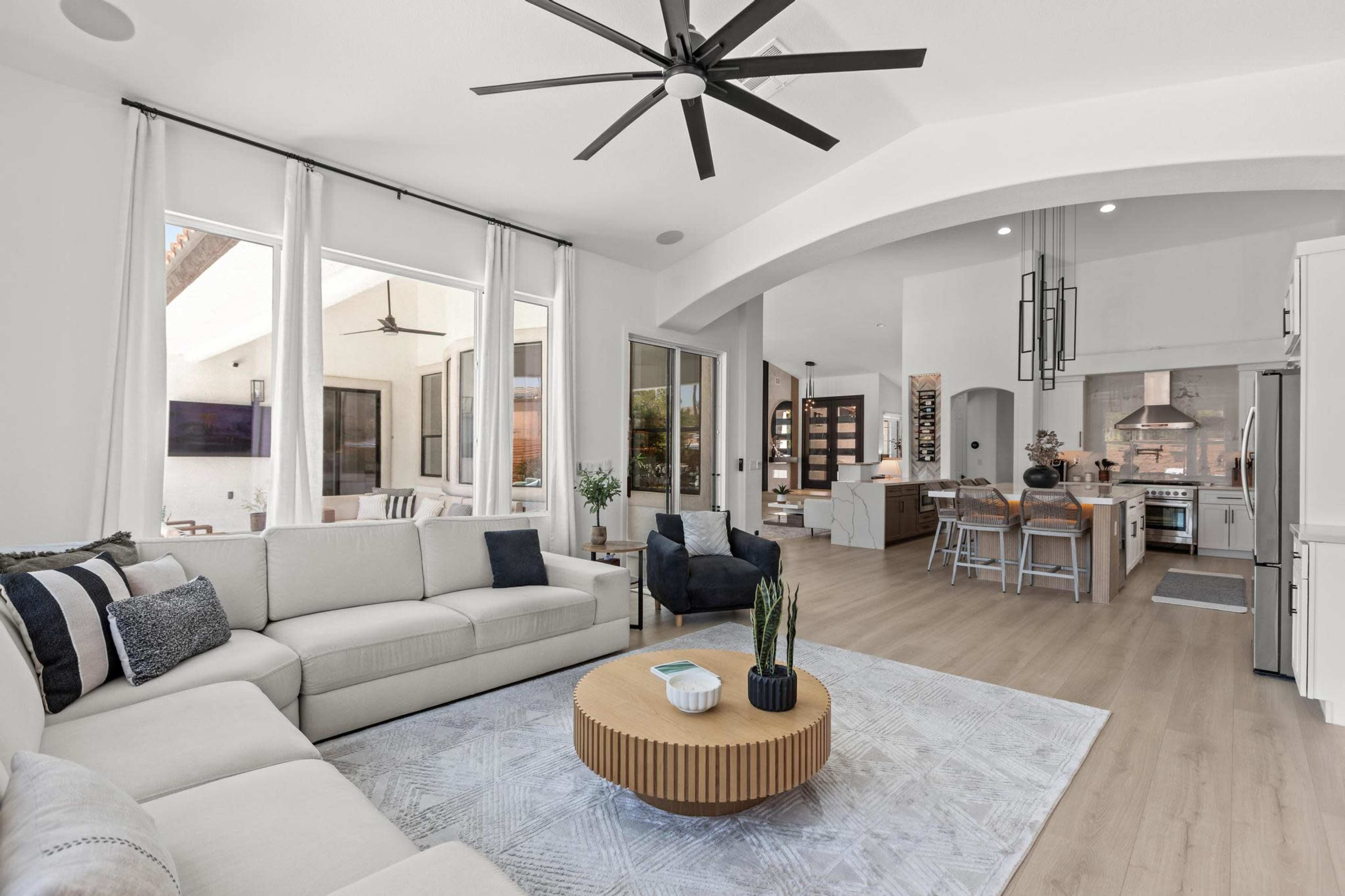 A spacious modern living room features a large sectional sofa, a round coffee table, and large windows that overlook an outdoor area, with an open kitchen visible in the background.