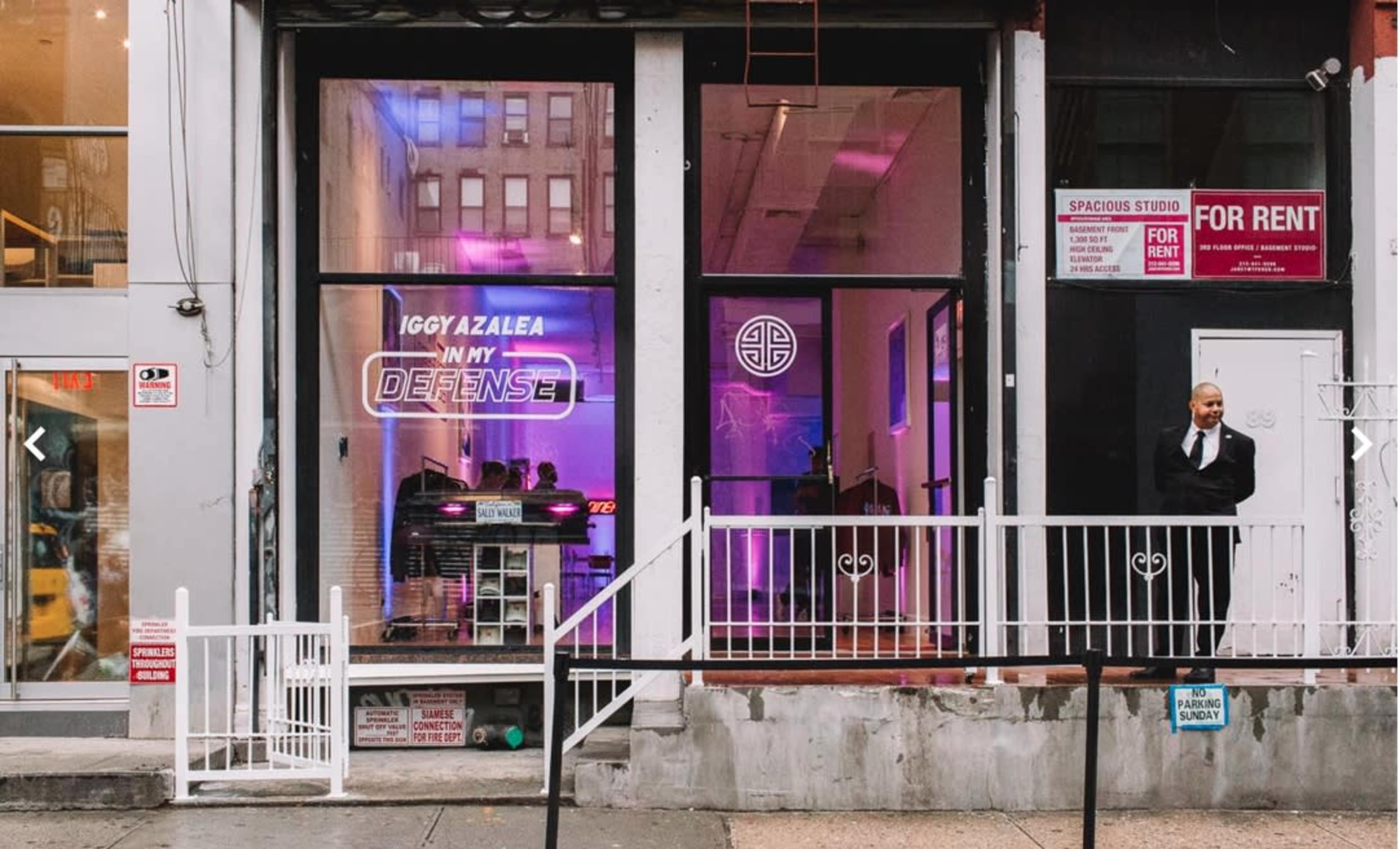 A storefront featuring neon signage that reads "IGGY AZALEA IN MY DEFENSE," with a security guard standing outside.