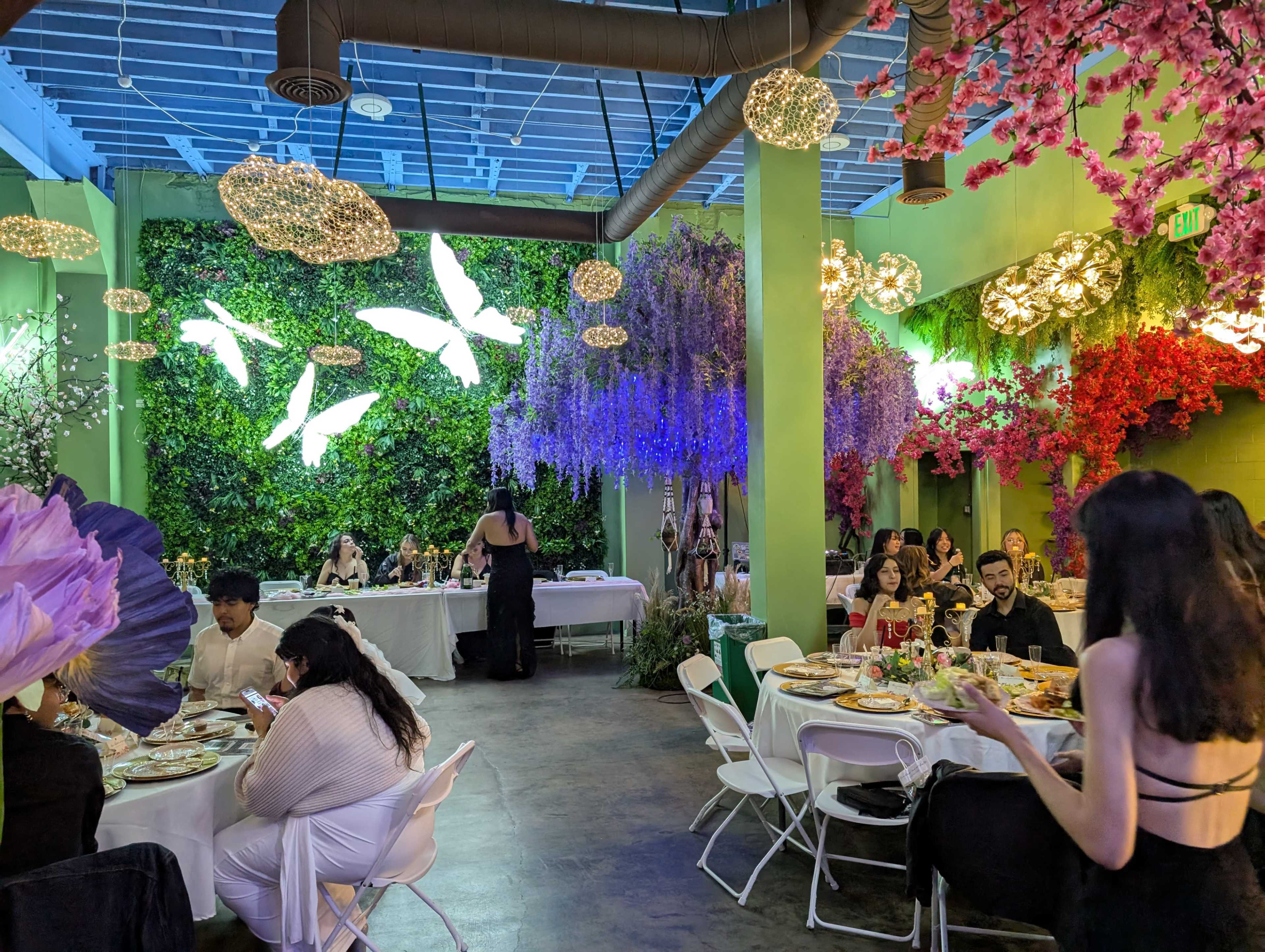 The scene features a vibrant dining area adorned with colorful floral decorations and illuminated by unique light fixtures, where guests are seated at elegantly set tables.