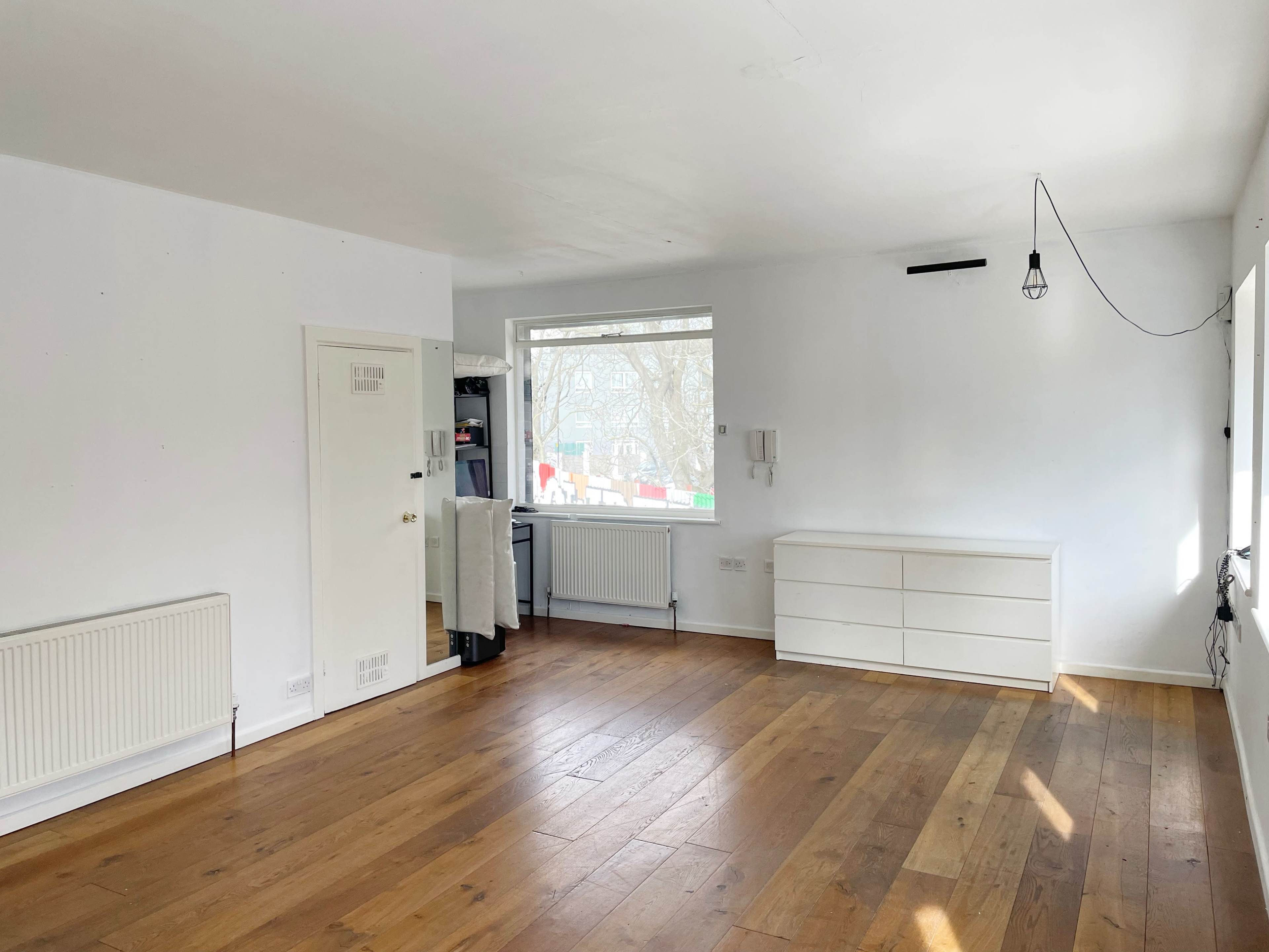 A spacious room with wooden flooring, a large window, a white dresser, and a light fixture hanging from the ceiling.