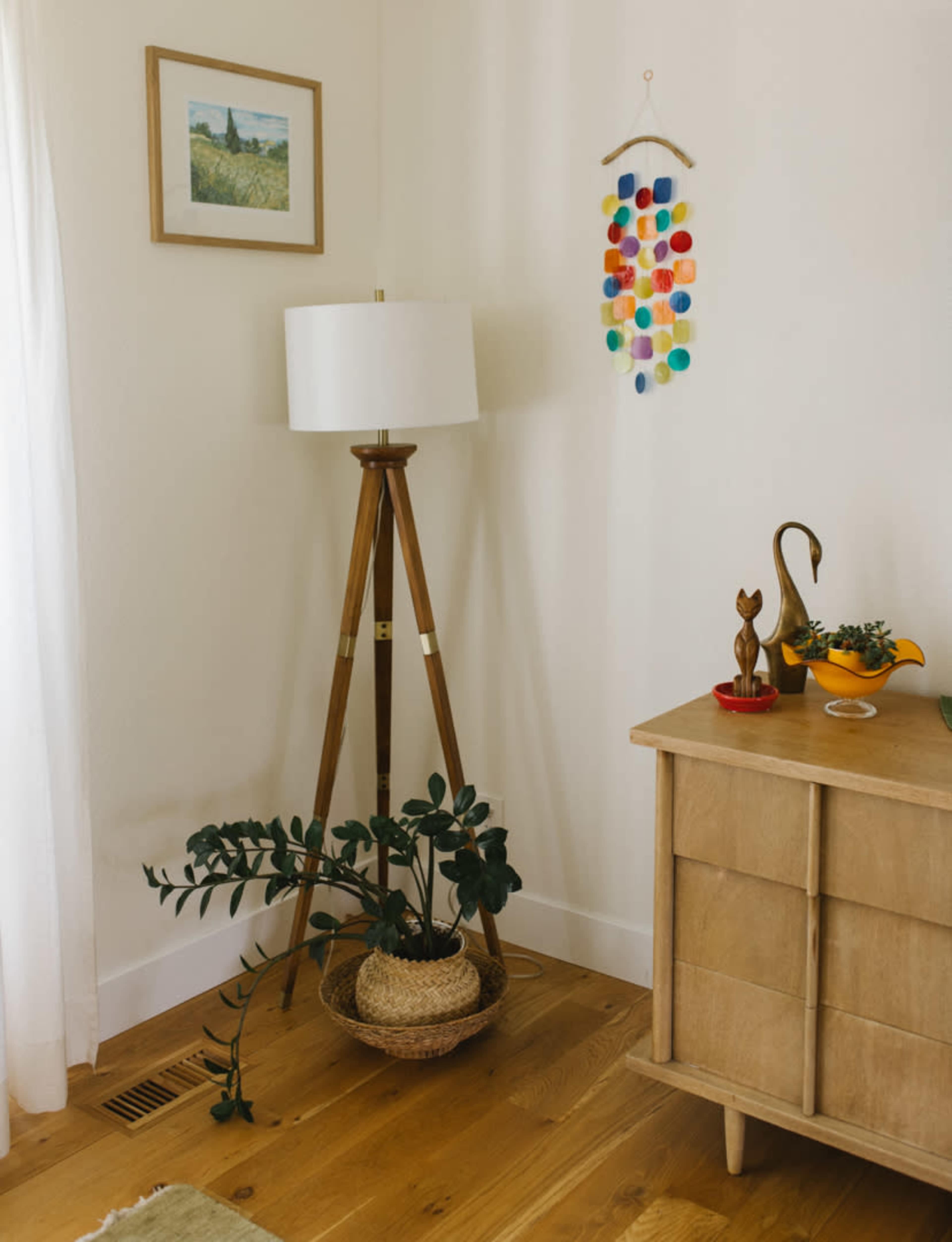 A corner of a room features a wooden tripod lamp, a colorful wall hanging, a small indoor plant in a woven basket, and a mid-century-style wooden dresser.