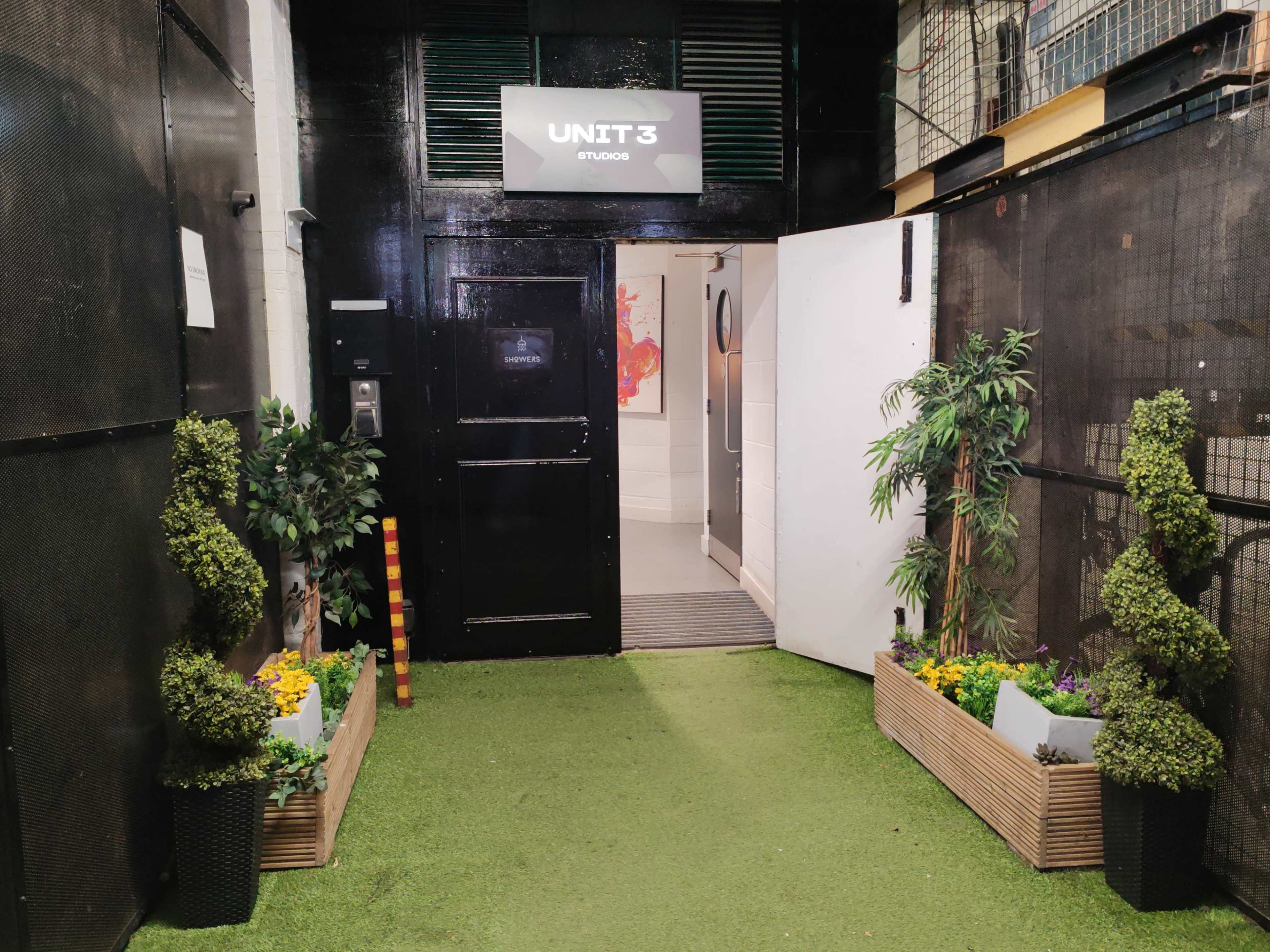 The image shows an entrance to a unit labeled "UNIT 3," flanked by planters with greenery and flowers on either side, leading to an interior space.