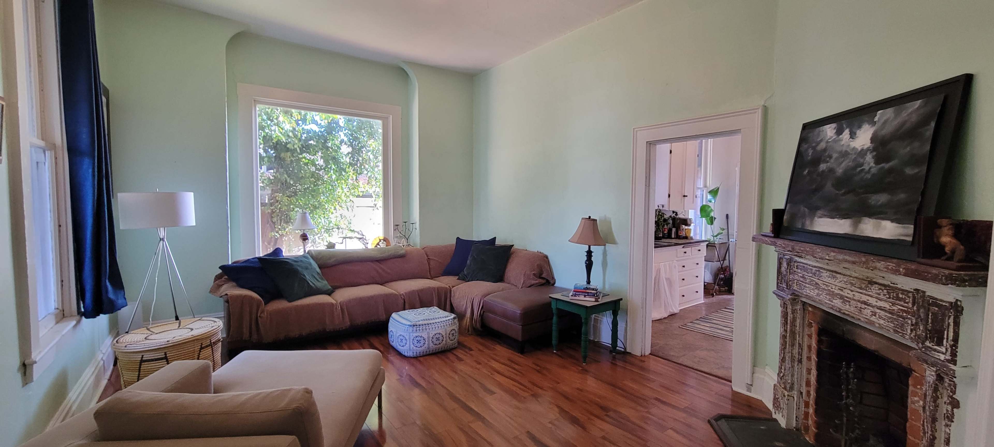 The image shows a comfortable living room with a sectional sofa, a lamp, a small table, and a fireplace, connected to a kitchen visible in the background.