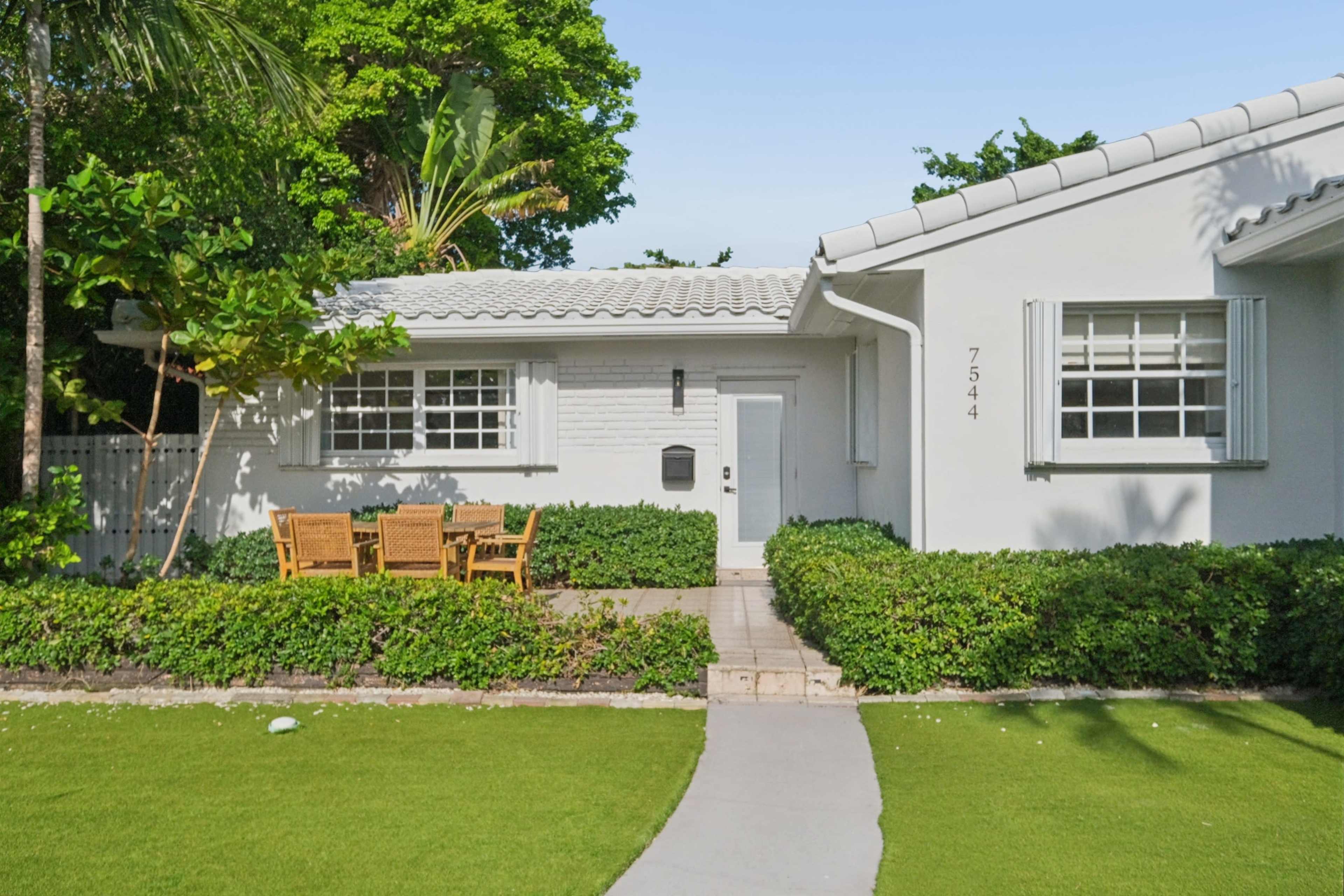 A white house with a tiled roof features a front patio with wooden furniture surrounded by green bushes and a neatly trimmed lawn.