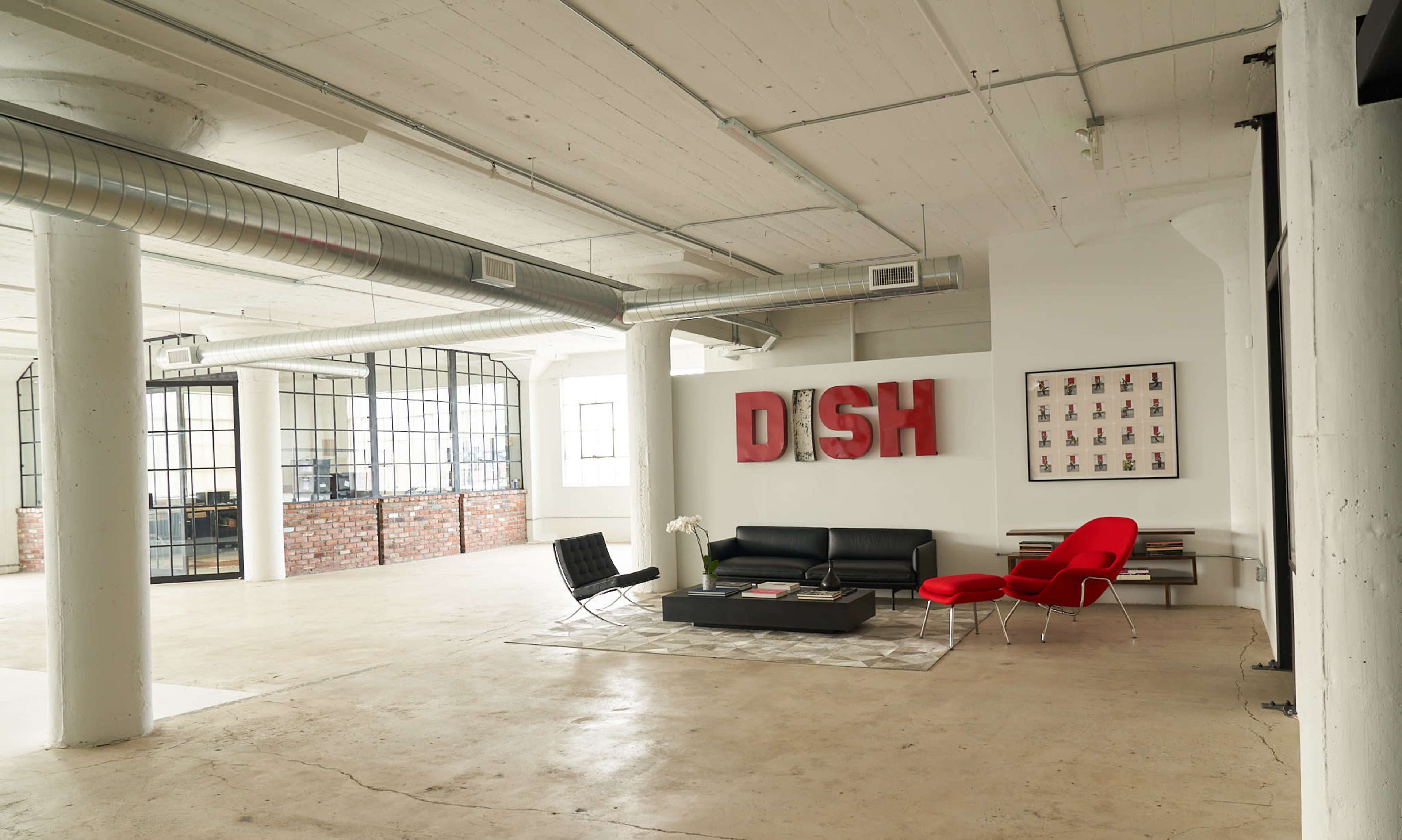 The image shows a spacious office interior with exposed concrete pillars, featuring modern furniture including a black sofa, a red chair, and a large decorative "DISH" sign on the wall.