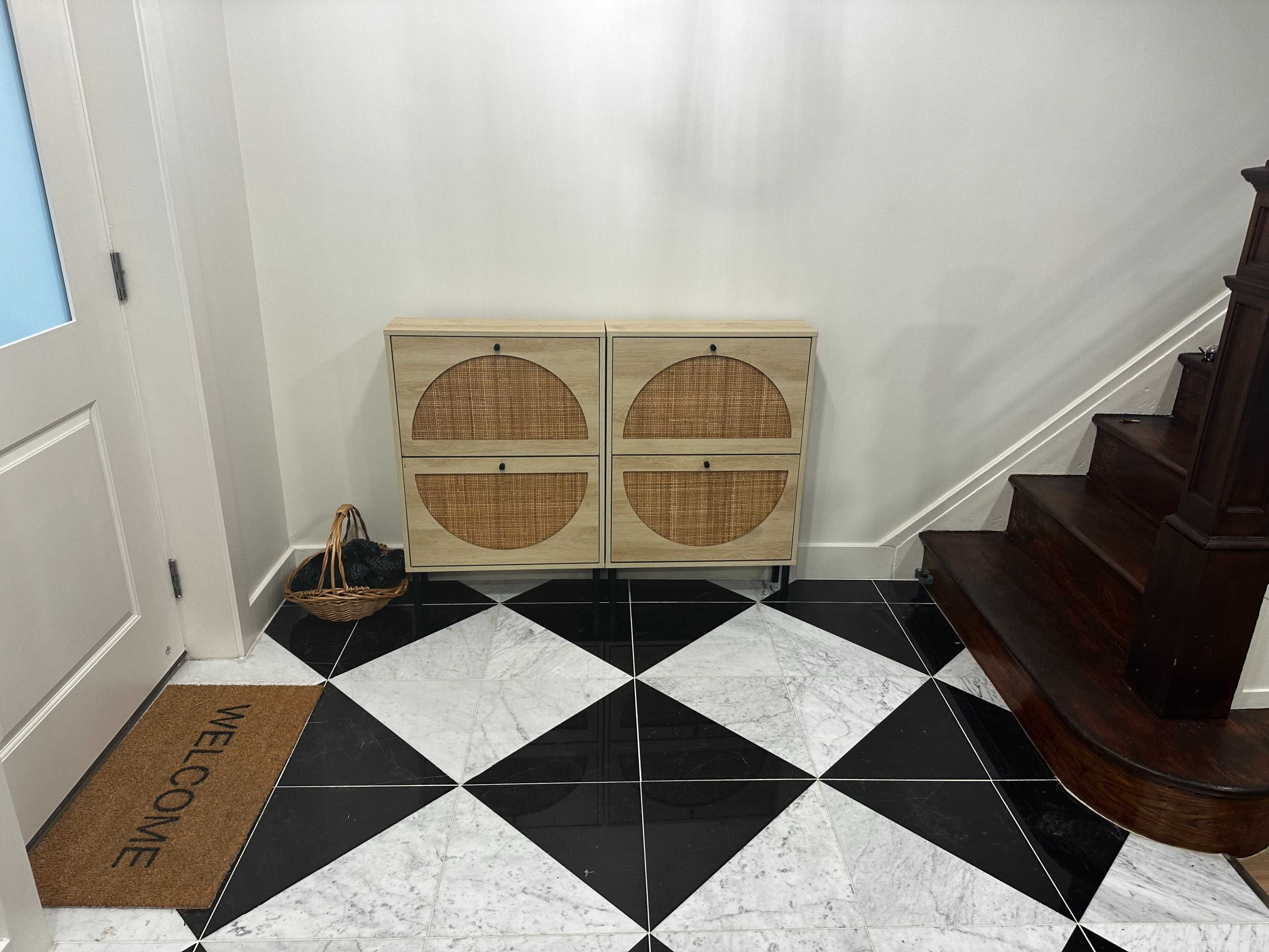 The image shows an entryway with a patterned black and white tiled floor, a wooden staircase, a cabinet with circular patterned doors, and a welcome mat.
