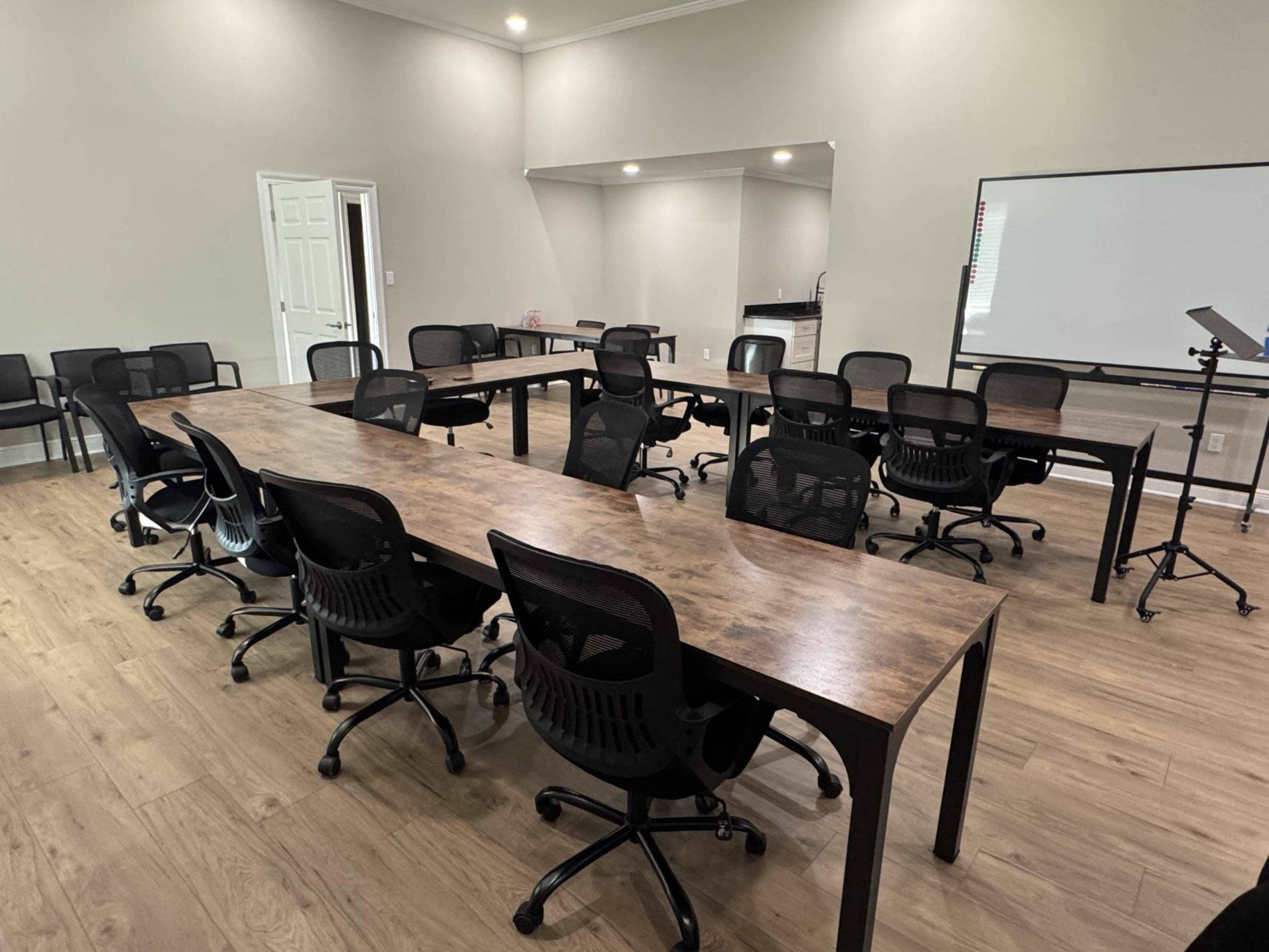 The image shows a spacious conference room arranged with several long tables and black office chairs, featuring a whiteboard and a kitchen area in the background.