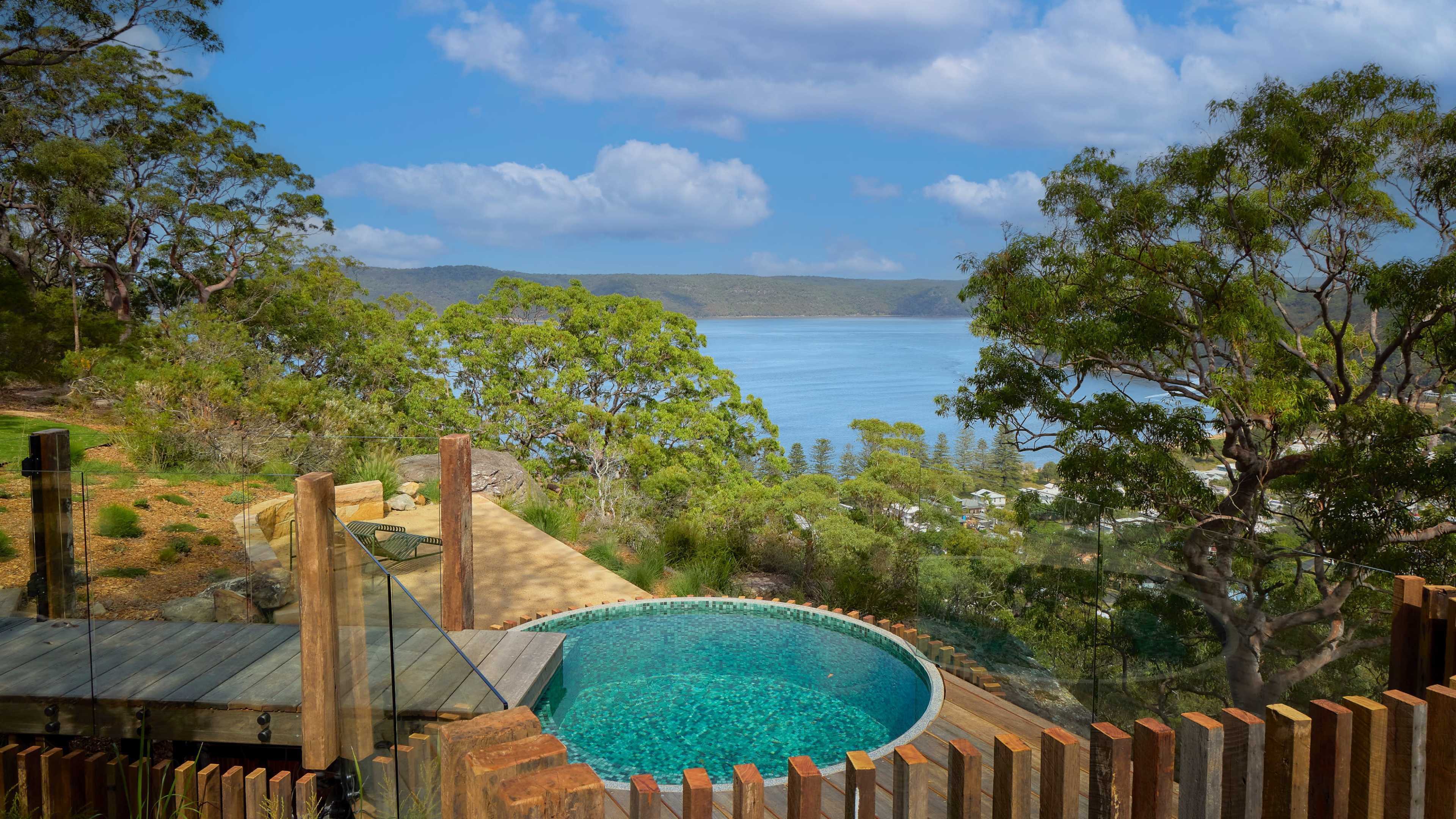 The image shows a circular pool overlooking a tranquil body of water and lush greenery in the background.