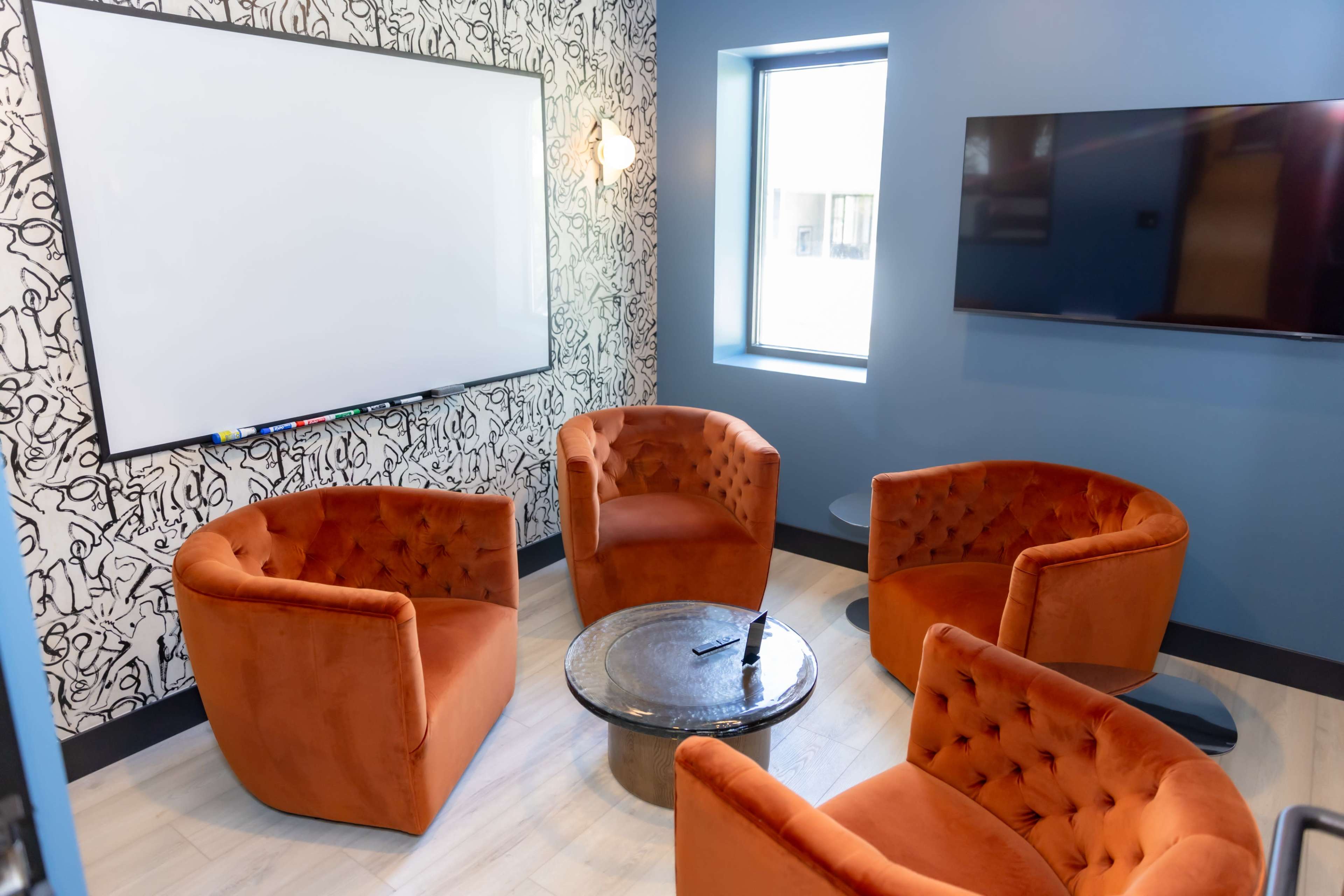 The image shows a small lounge area with four orange upholstered chairs arranged around a round table, a whiteboard on one wall, and a wall-mounted TV on another.