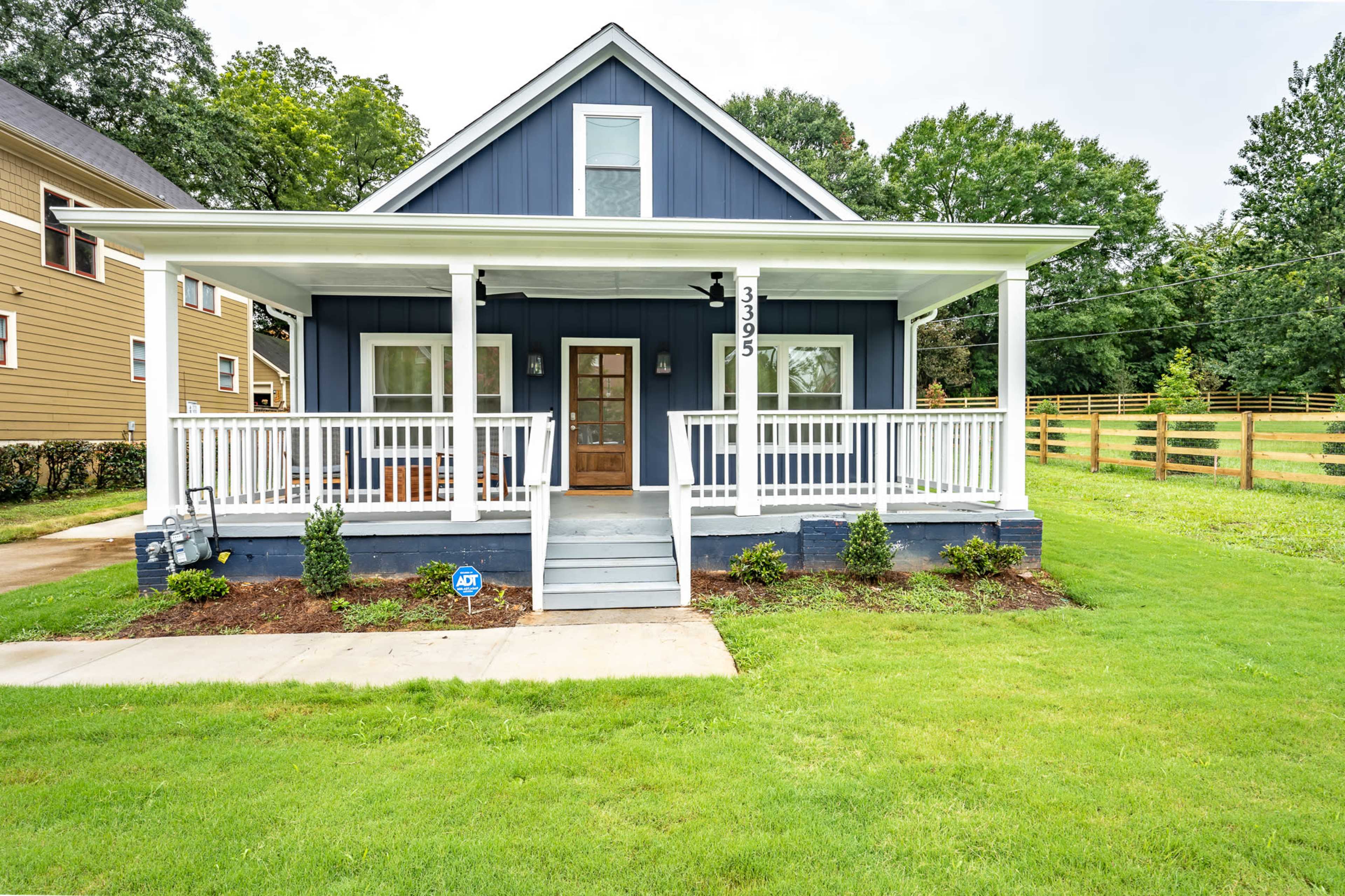A blue two-story house with a front porch and white railings is set on a grassy lawn with landscaping.