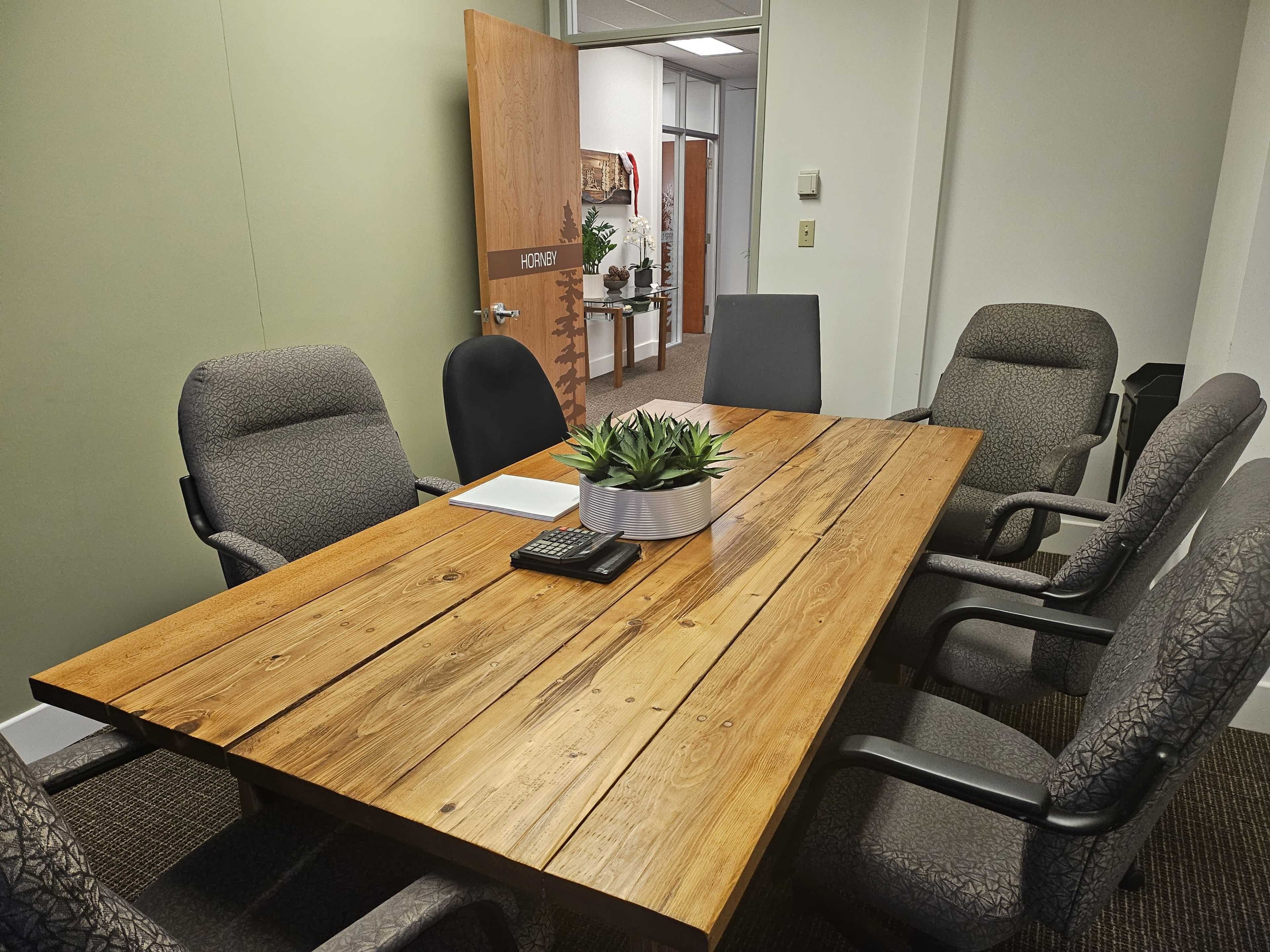 A wooden conference table surrounded by six chairs is situated in a meeting room, with a potted plant and a notepad on the table.