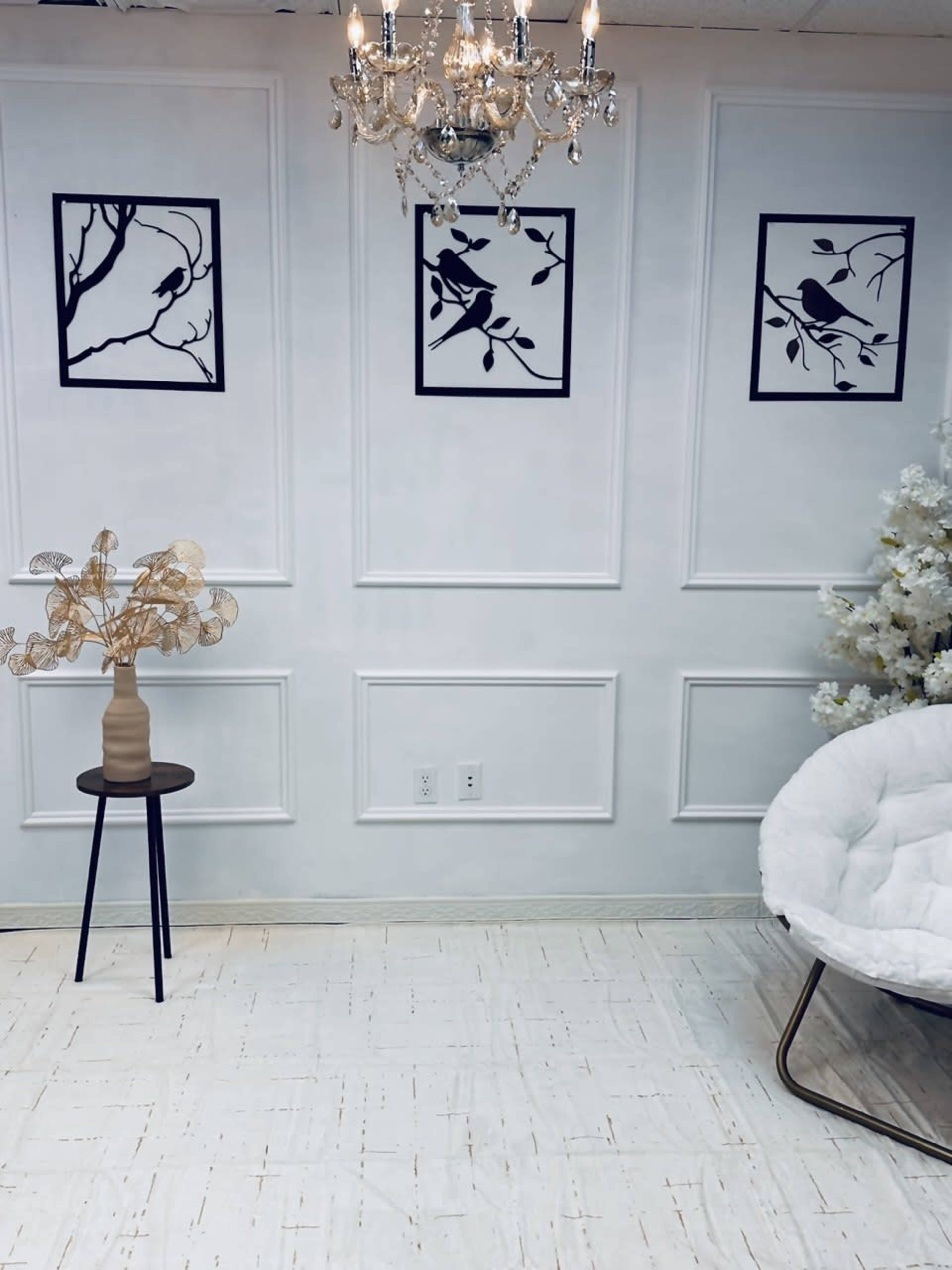 The image shows a neatly arranged room with white walls featuring framed black and white bird silhouettes, a small table with a vase, and a plush chair.