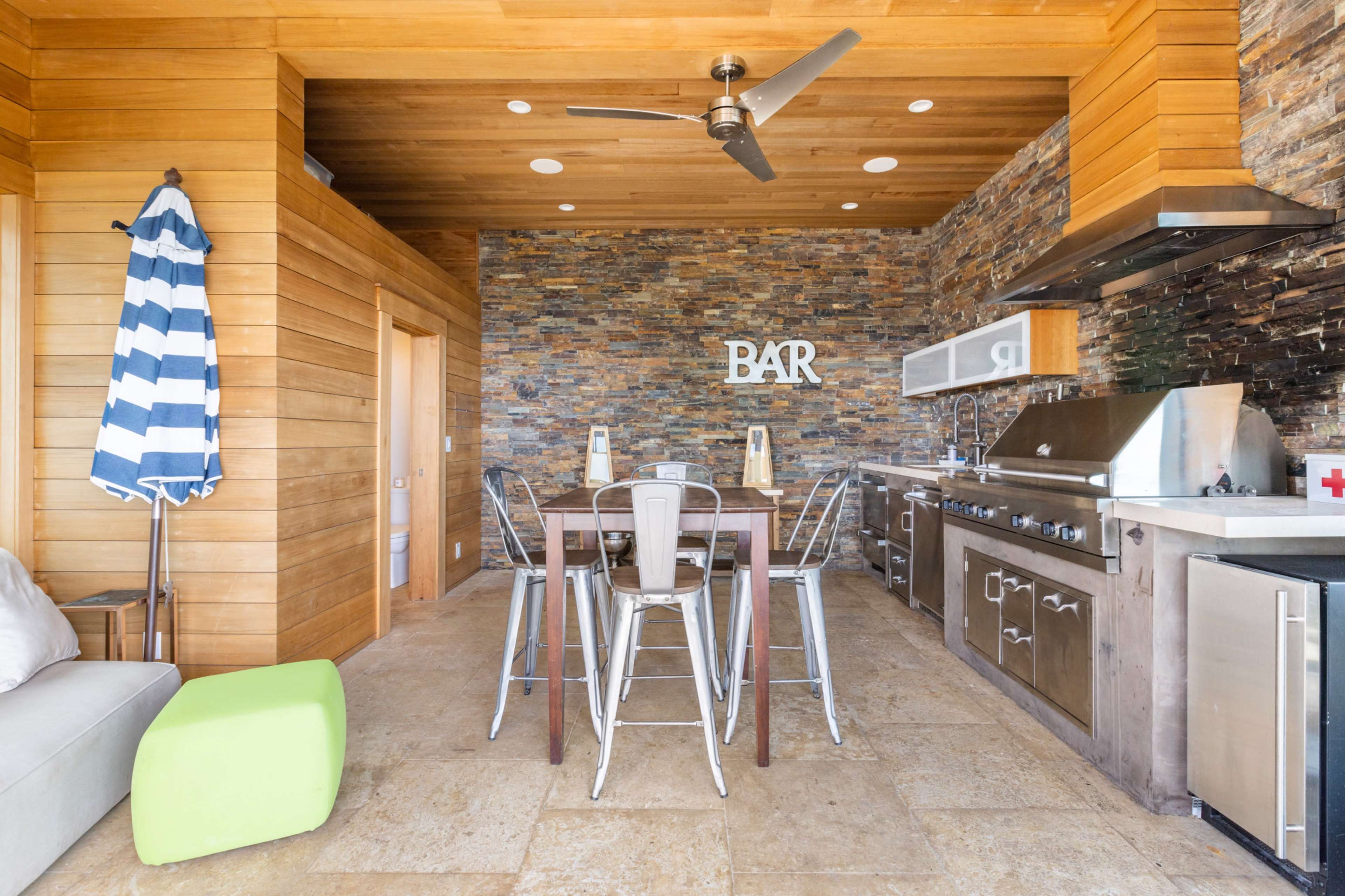 An outdoor kitchen and bar area with a wooden wall, stone accents, stainless steel appliances, and seating.