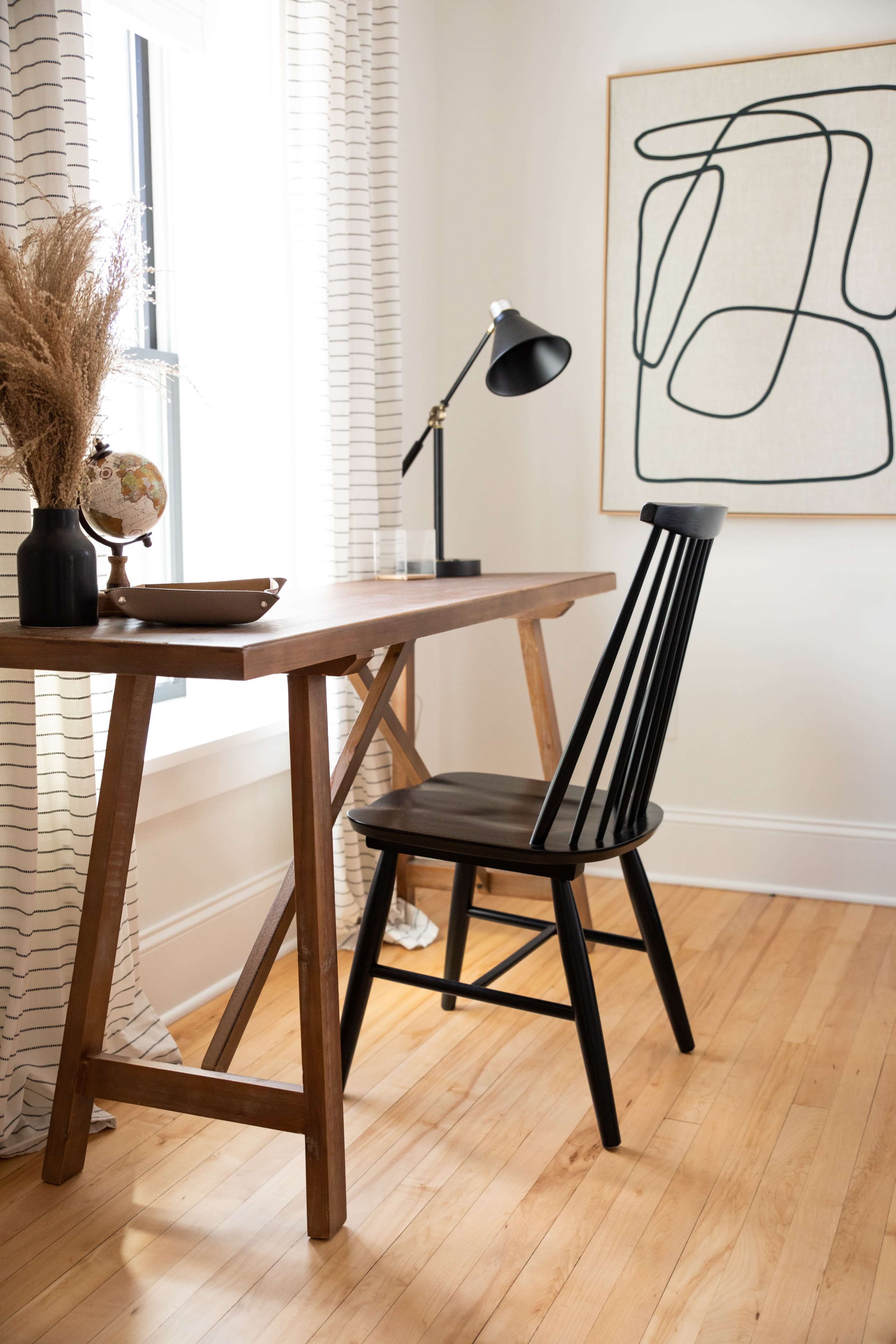 A wooden desk with a black chair is positioned beside a window, accompanied by a globe and a modern lamp, with a large line art print hanging on the wall.