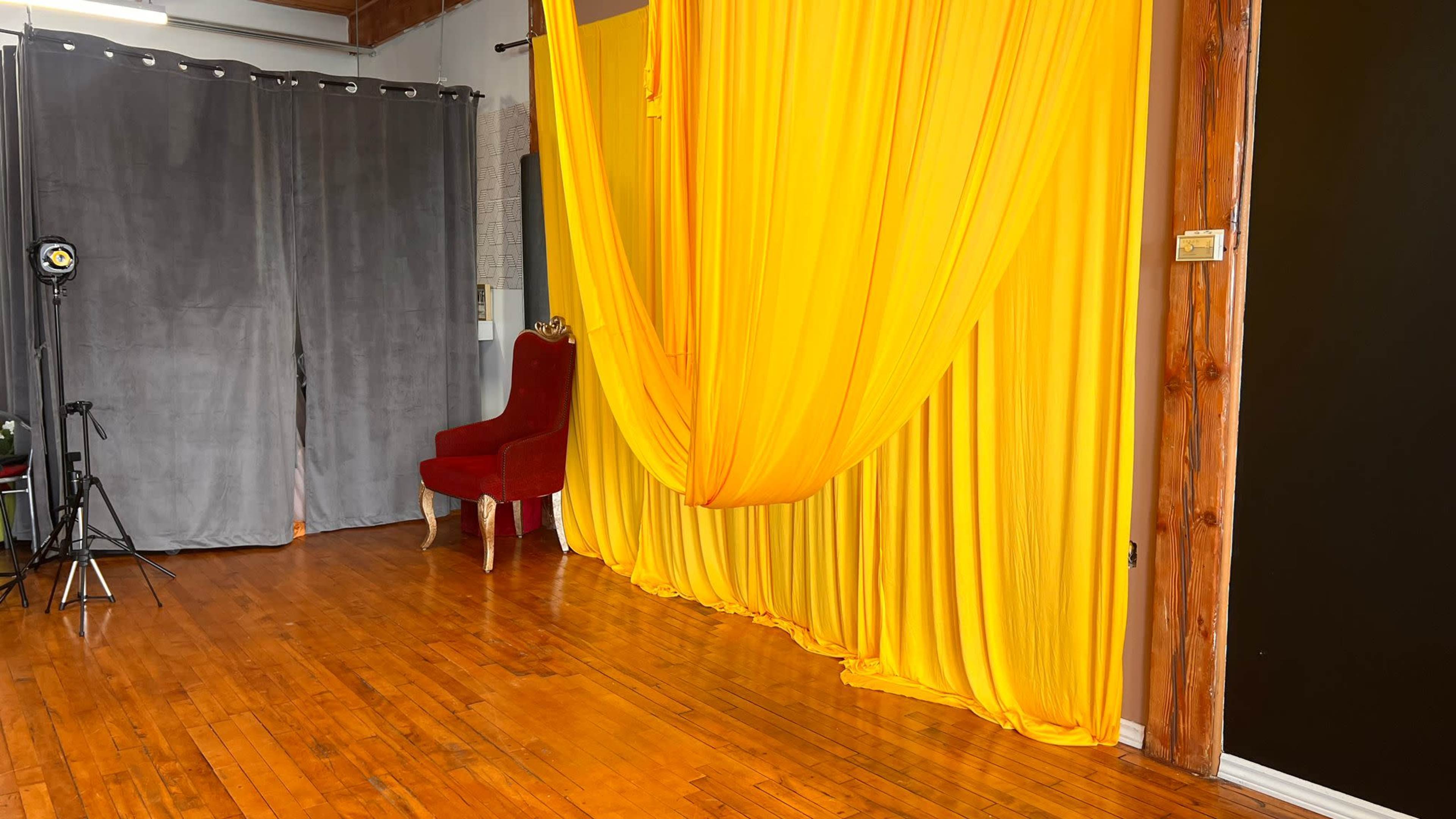 A spacious room with a yellow draped backdrop, a red velvet chair, and gray curtains, all set on polished wooden floors.