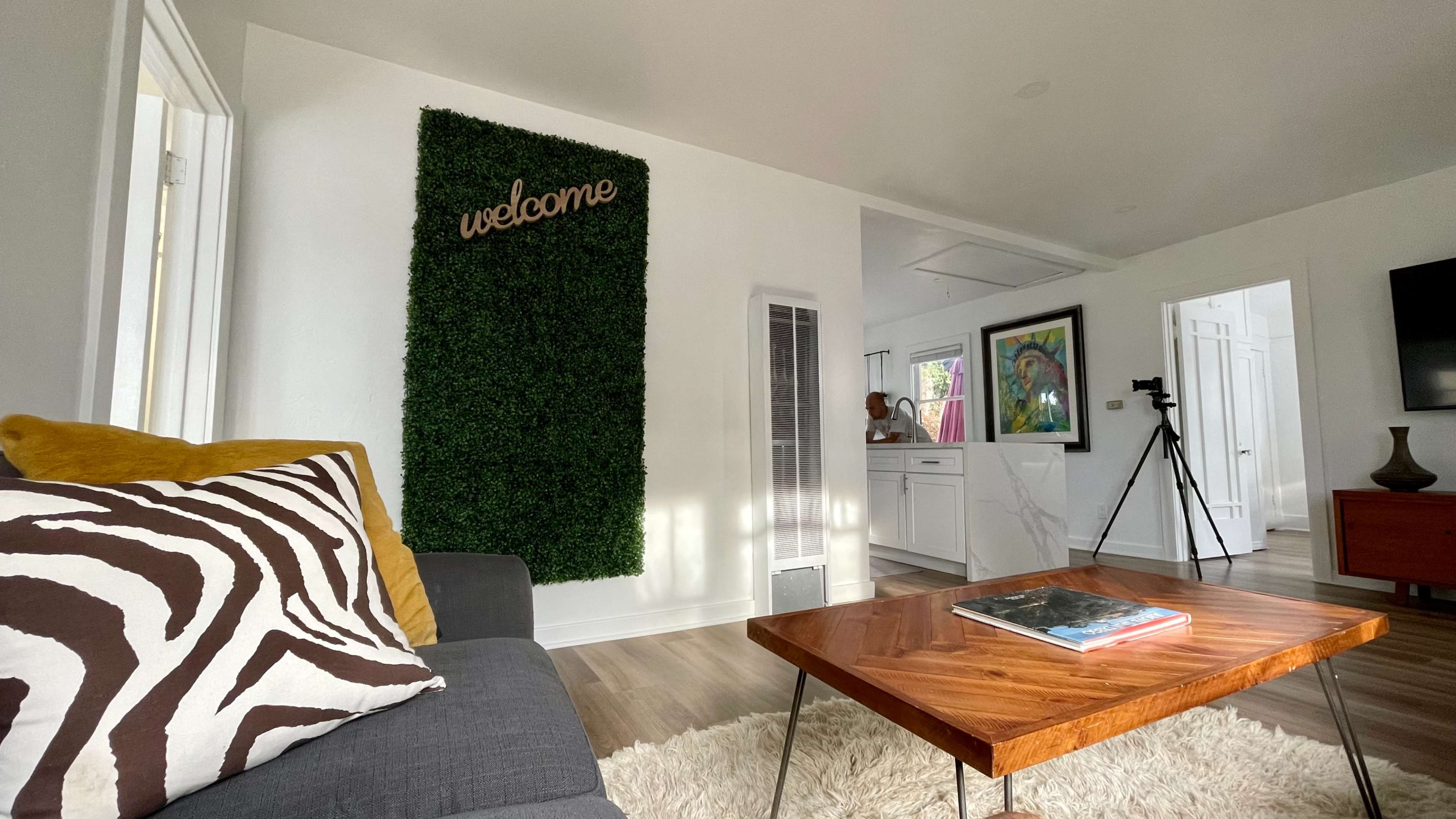 The living room features a sofa with a zebra-striped cushion, a wooden coffee table, a welcome sign against a green wall, and a view of the kitchen area.