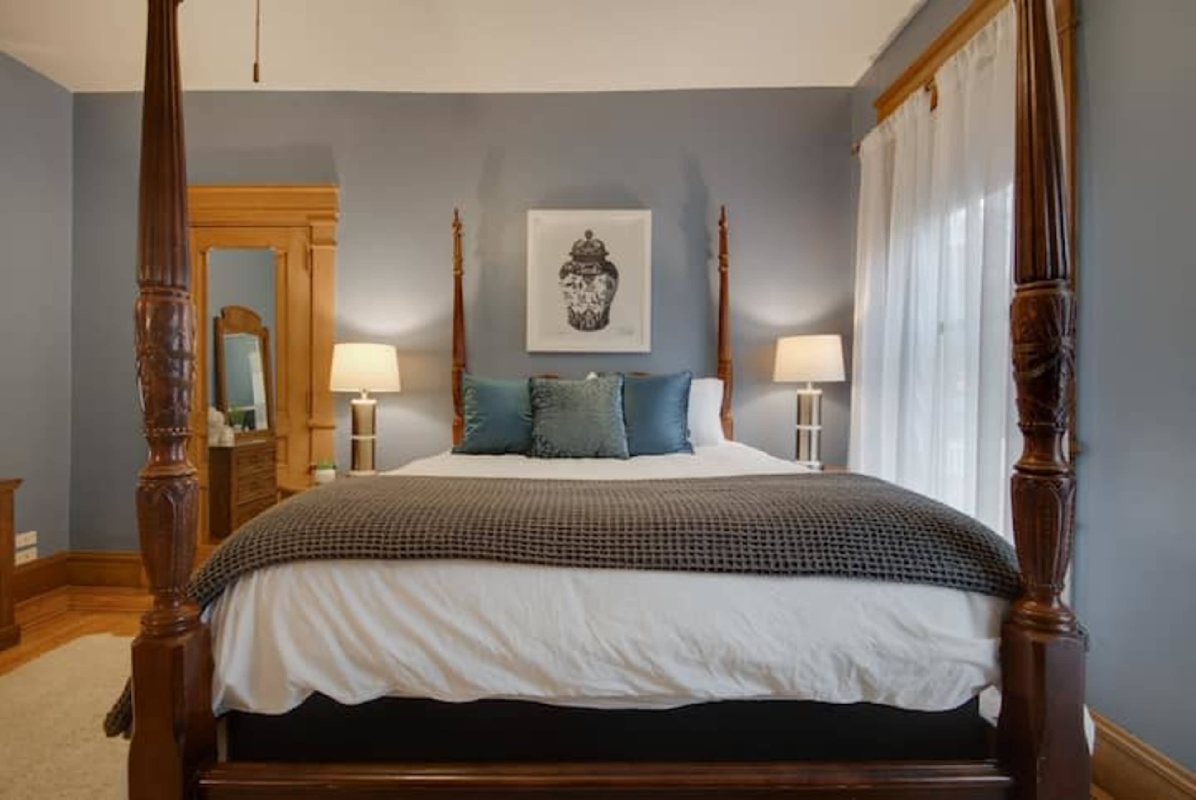 A spacious bedroom featuring a four-poster bed with a neutral comforter and blue throw pillows, flanked by two bedside lamps.