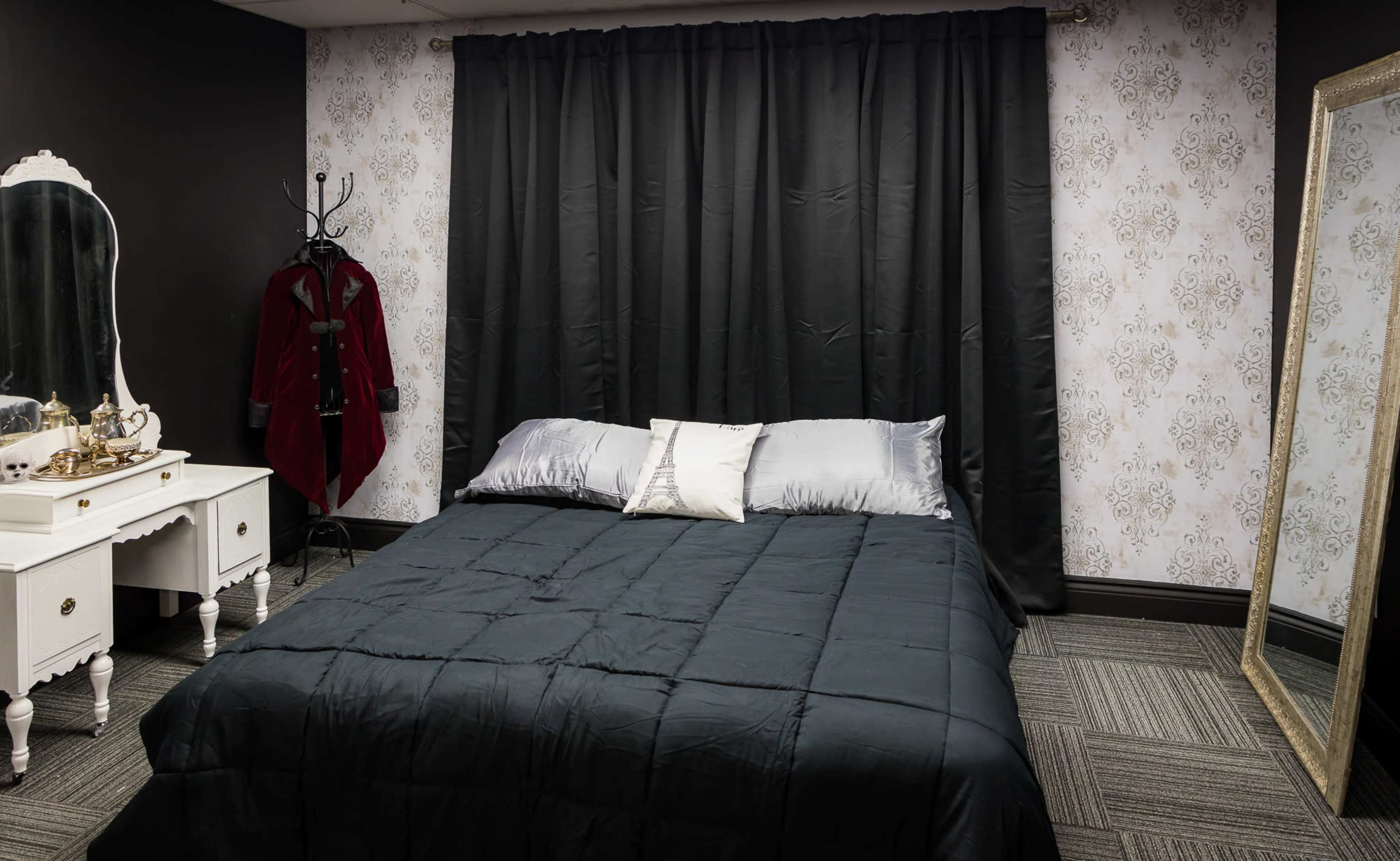 A neatly made bed with a dark comforter is positioned in a room featuring a black and white wallpapered wall, along with a large mirror and a white dresser.