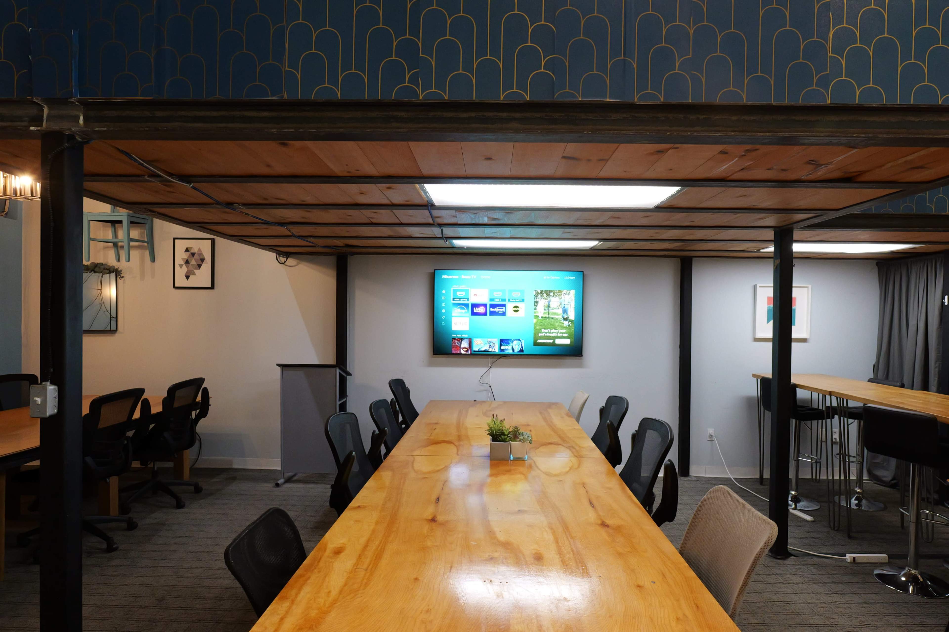 The image shows a modern conference room with a long wooden table, several ergonomic chairs, and a wall-mounted screen displaying a presentation.