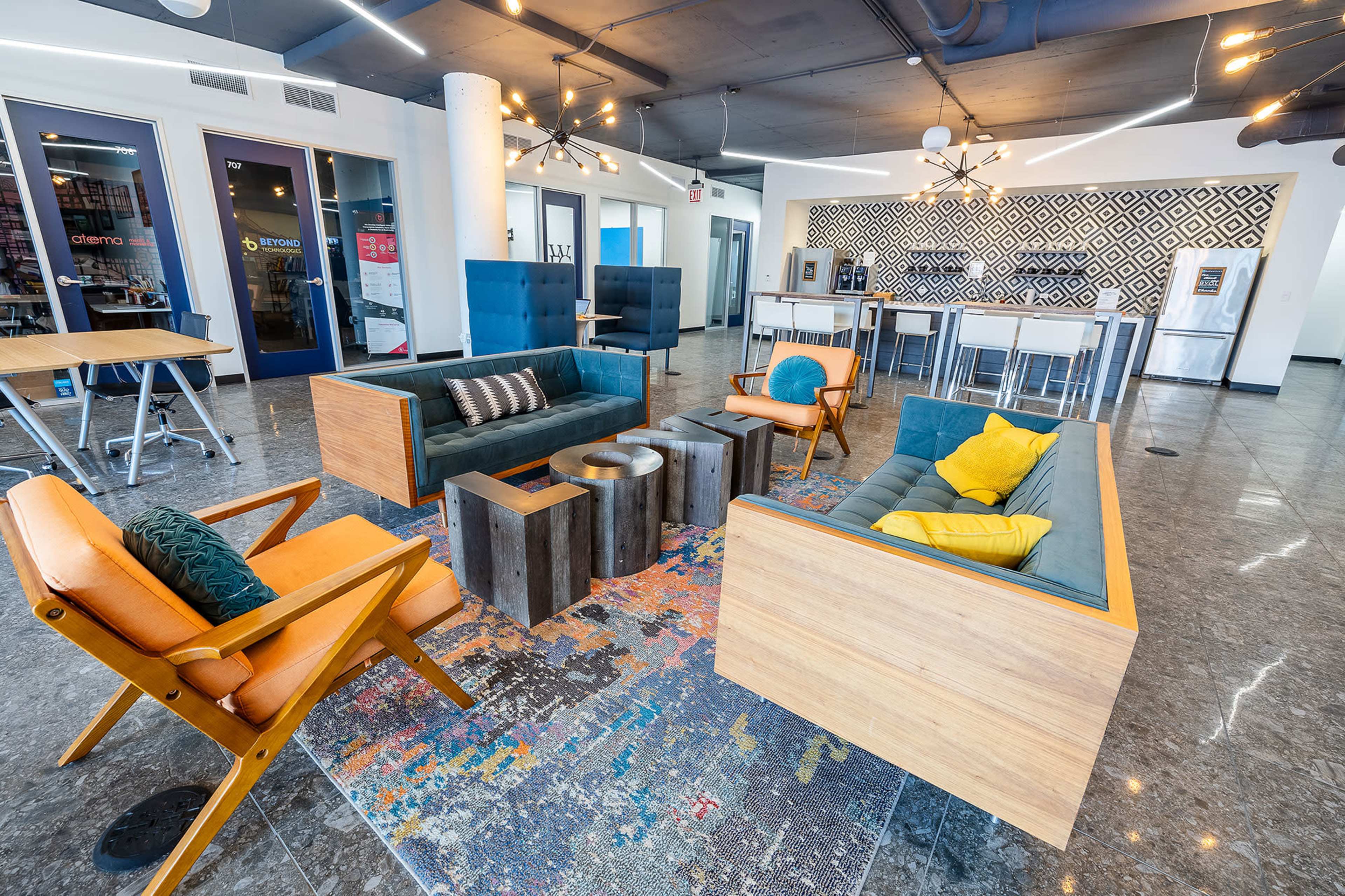 The scene features a modern lounge area with two wooden-framed sofas, a colorful area rug, and a bar-style kitchen in the background.