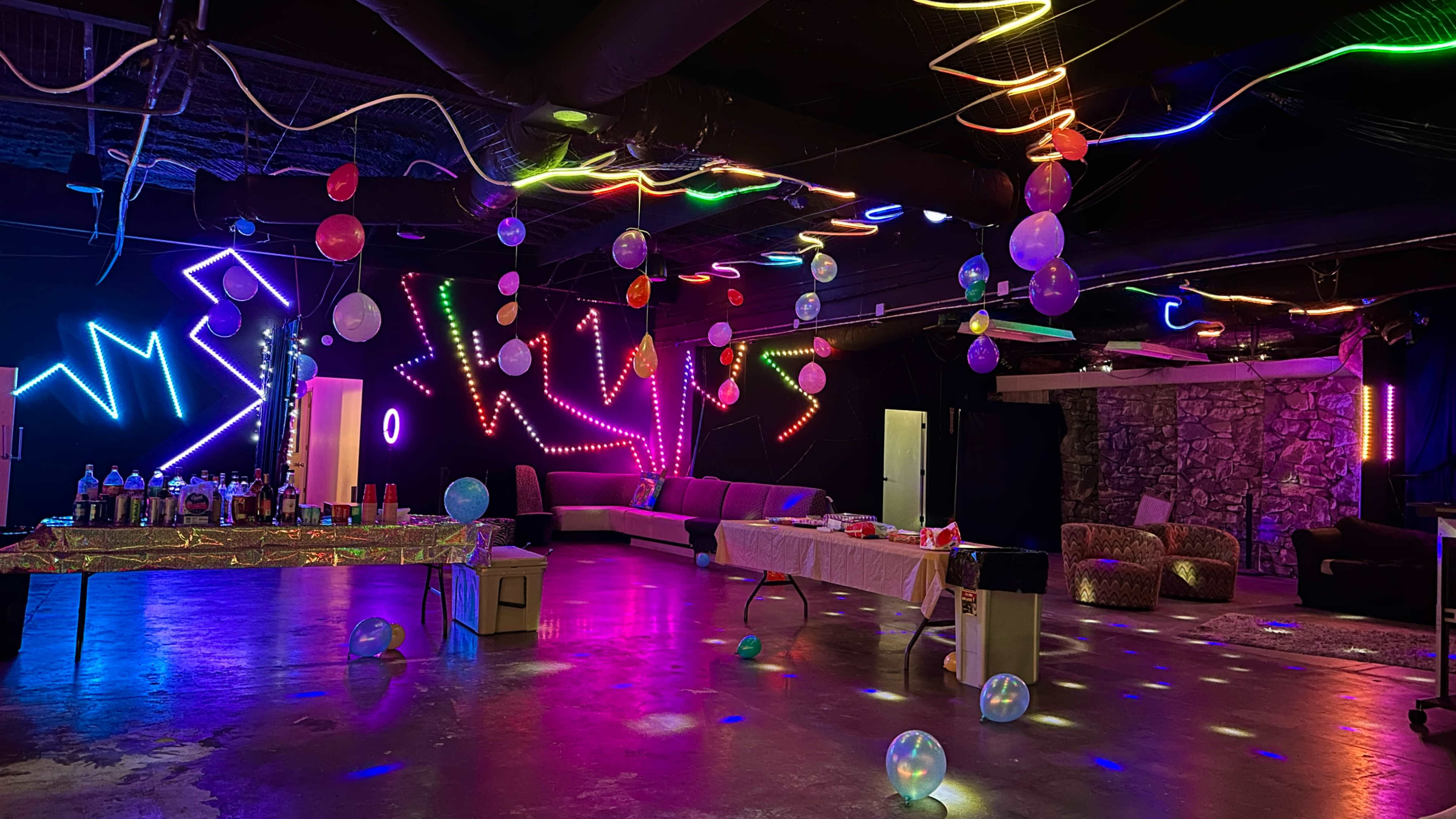 A brightly colored party space features glowing lights, balloons hanging from the ceiling, and tables set up with food and drinks.