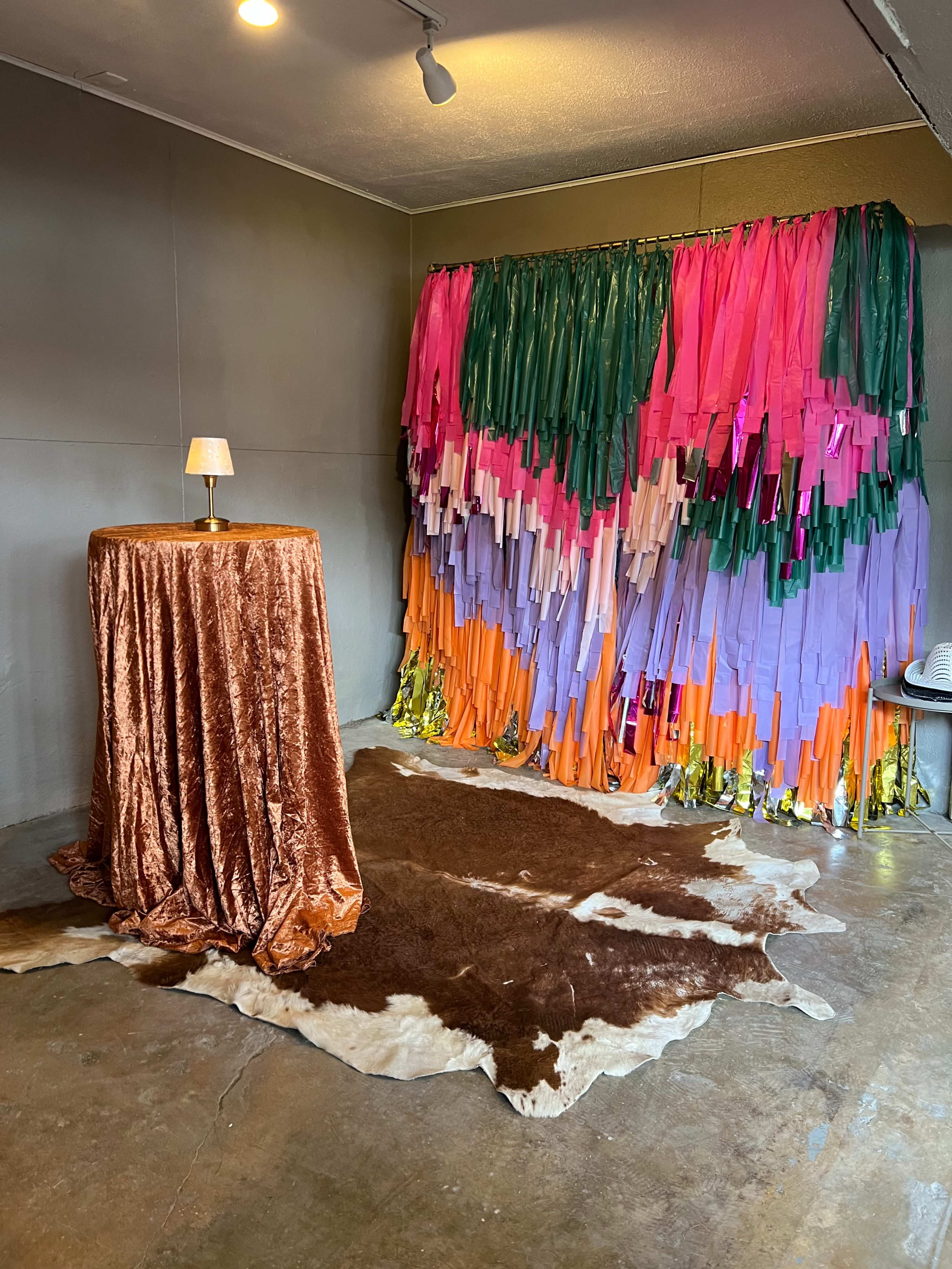 The image shows a small room with a colorful backdrop made of fabric strips, a brown table covered with a velvet cloth, and a cowhide rug on the floor.
