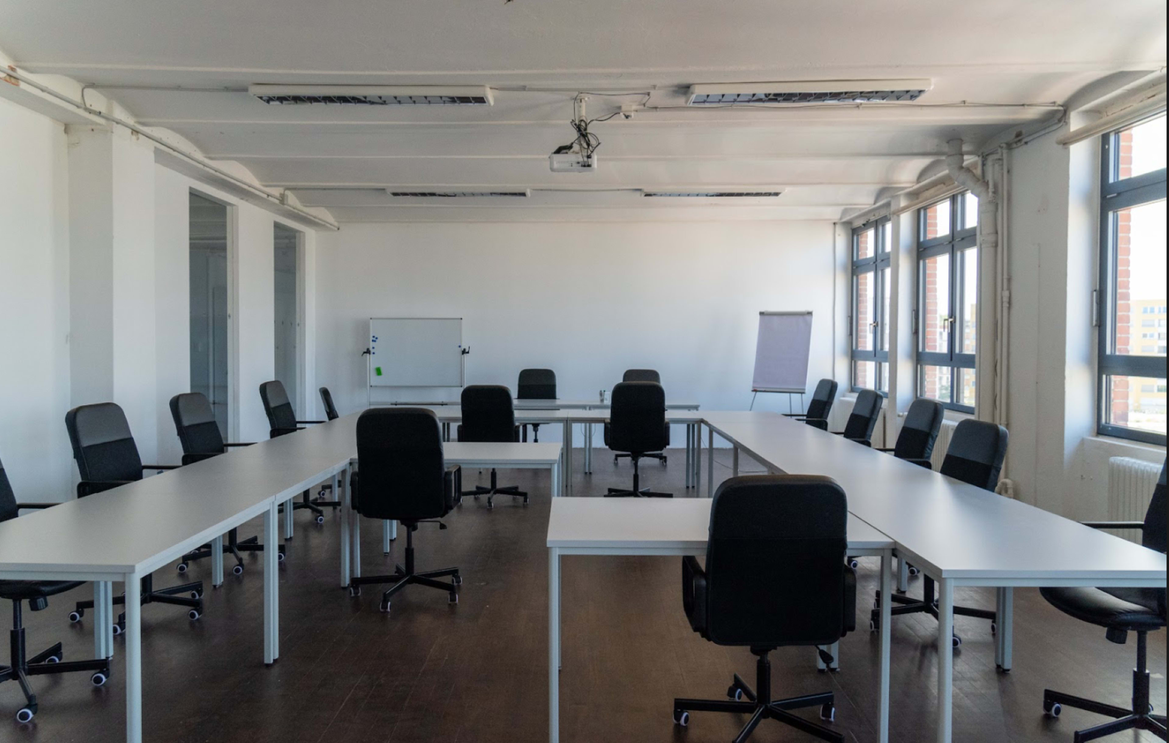 A large, empty conference room features a U-shaped arrangement of white tables and black rolling chairs under natural light from multiple windows.