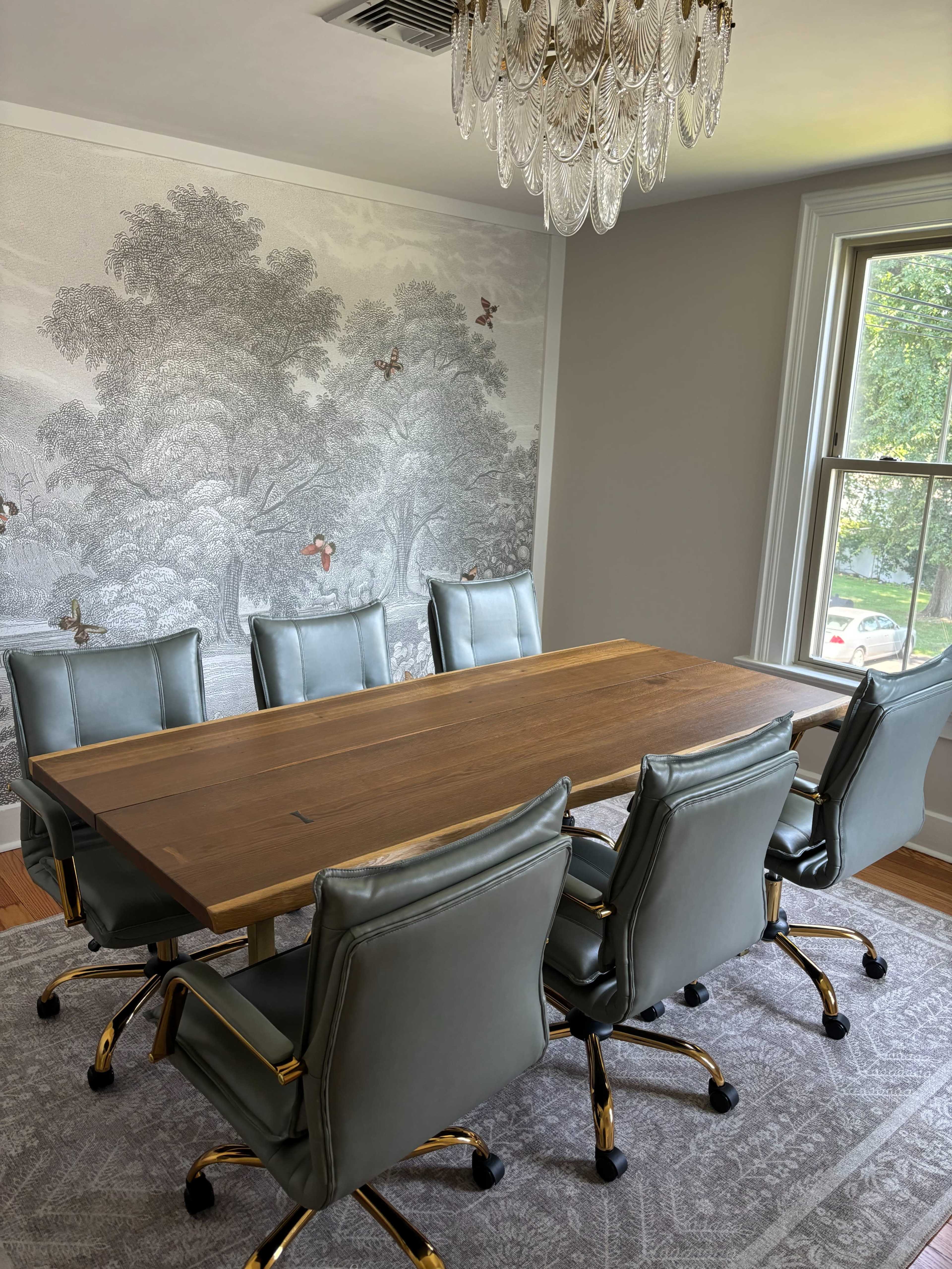 The image showcases a modern conference room with a long wooden table surrounded by six gray office chairs, set against a decorative wallpaper featuring a tree design.
