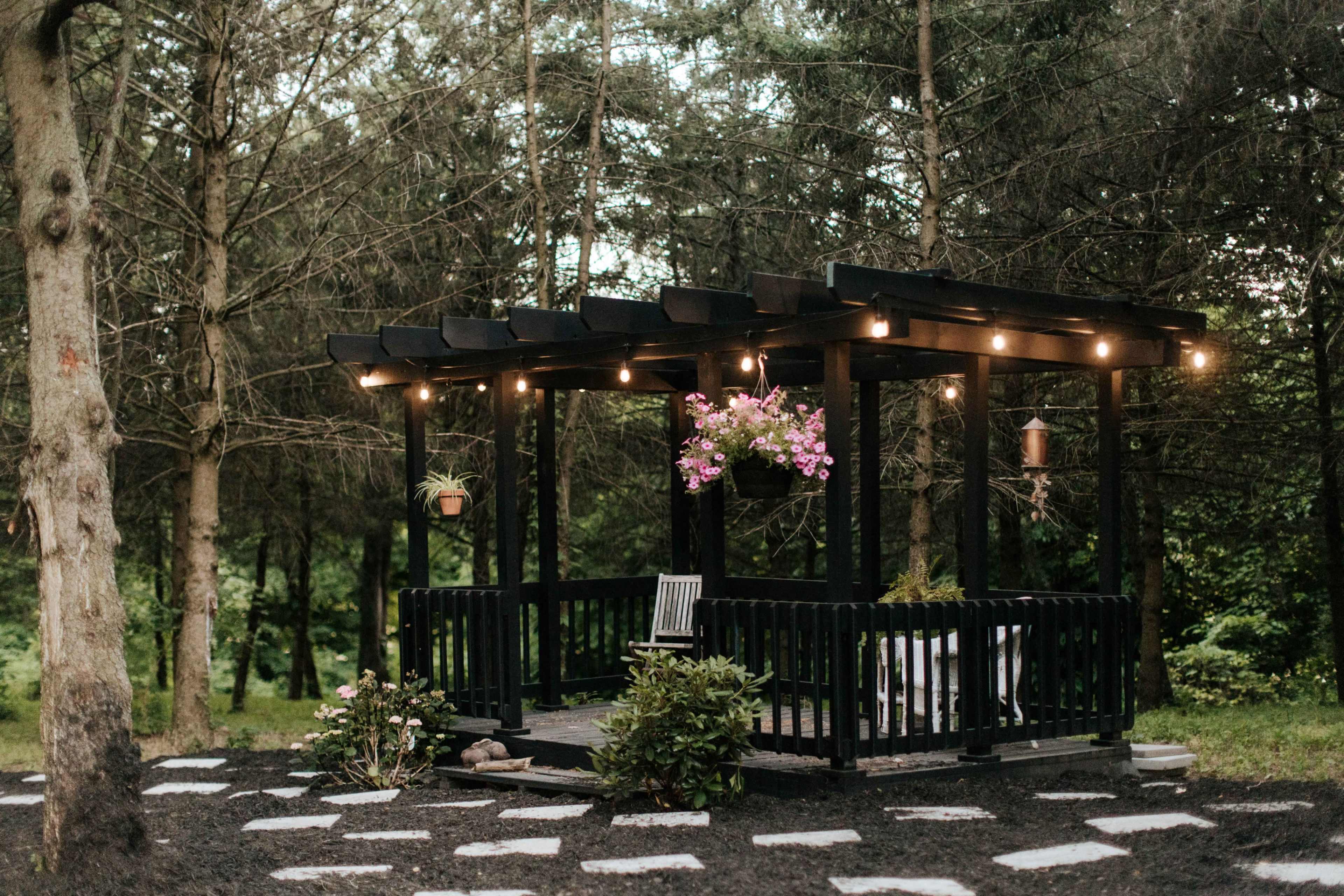 A black wooden gazebo with hanging plants and string lights is situated among trees in a garden area with stone paths.