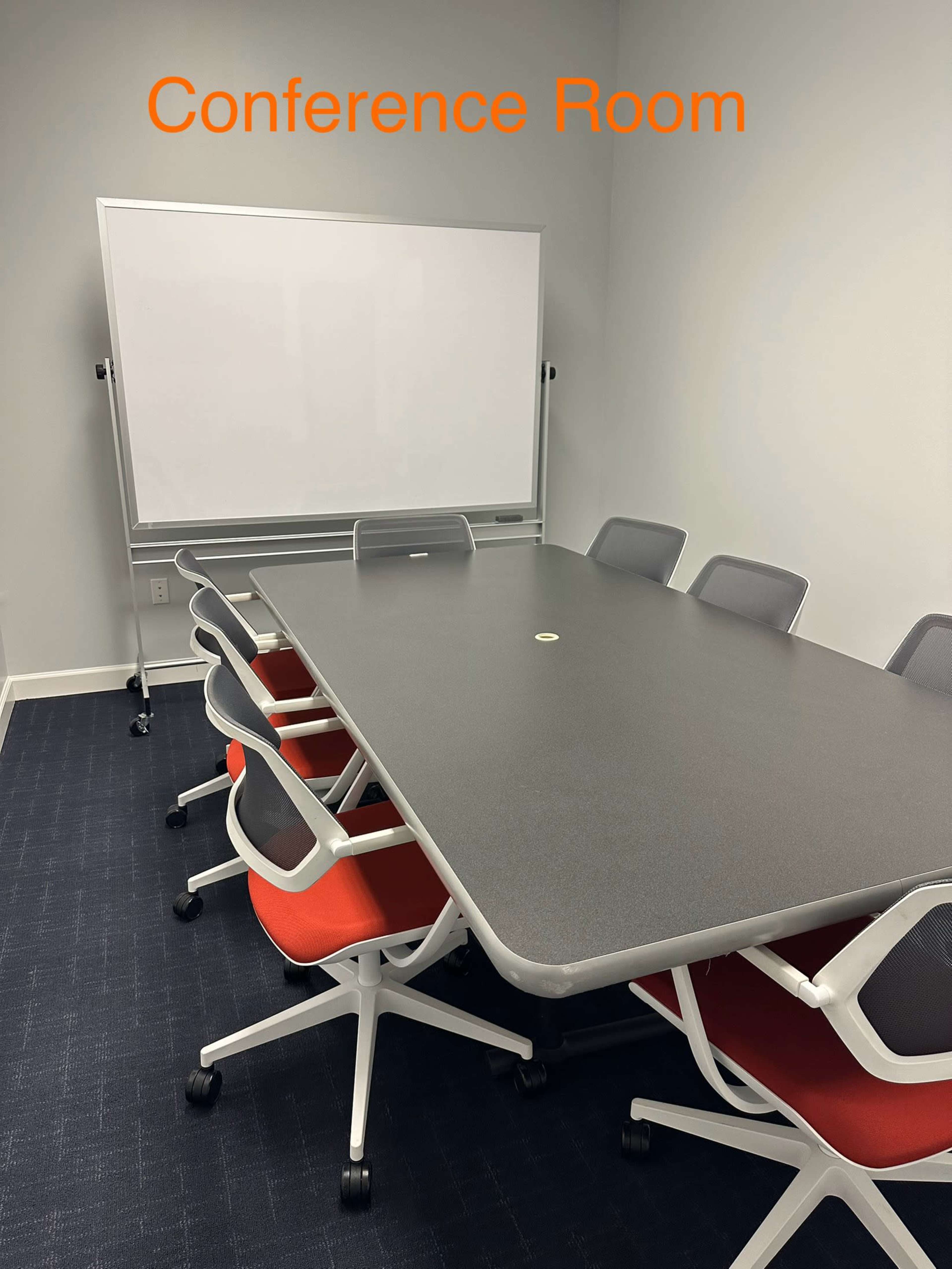 A conference room features a long gray table with eight chairs, each with a red seat, and a mounted whiteboard on a nearby wall.