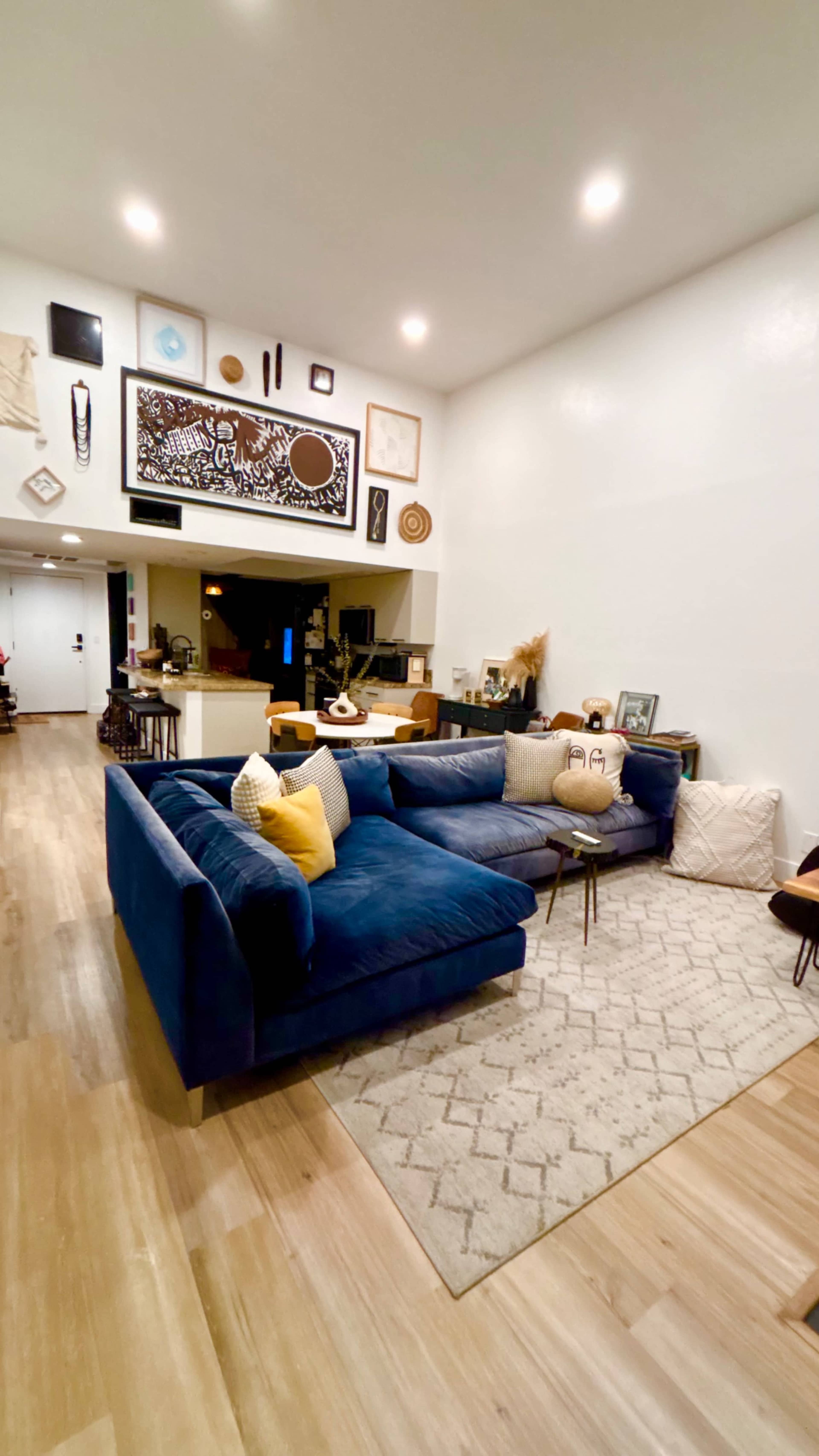 The image shows a spacious living room with a blue sectional couch, a light-colored rug, and various decorative art pieces displayed on the walls.