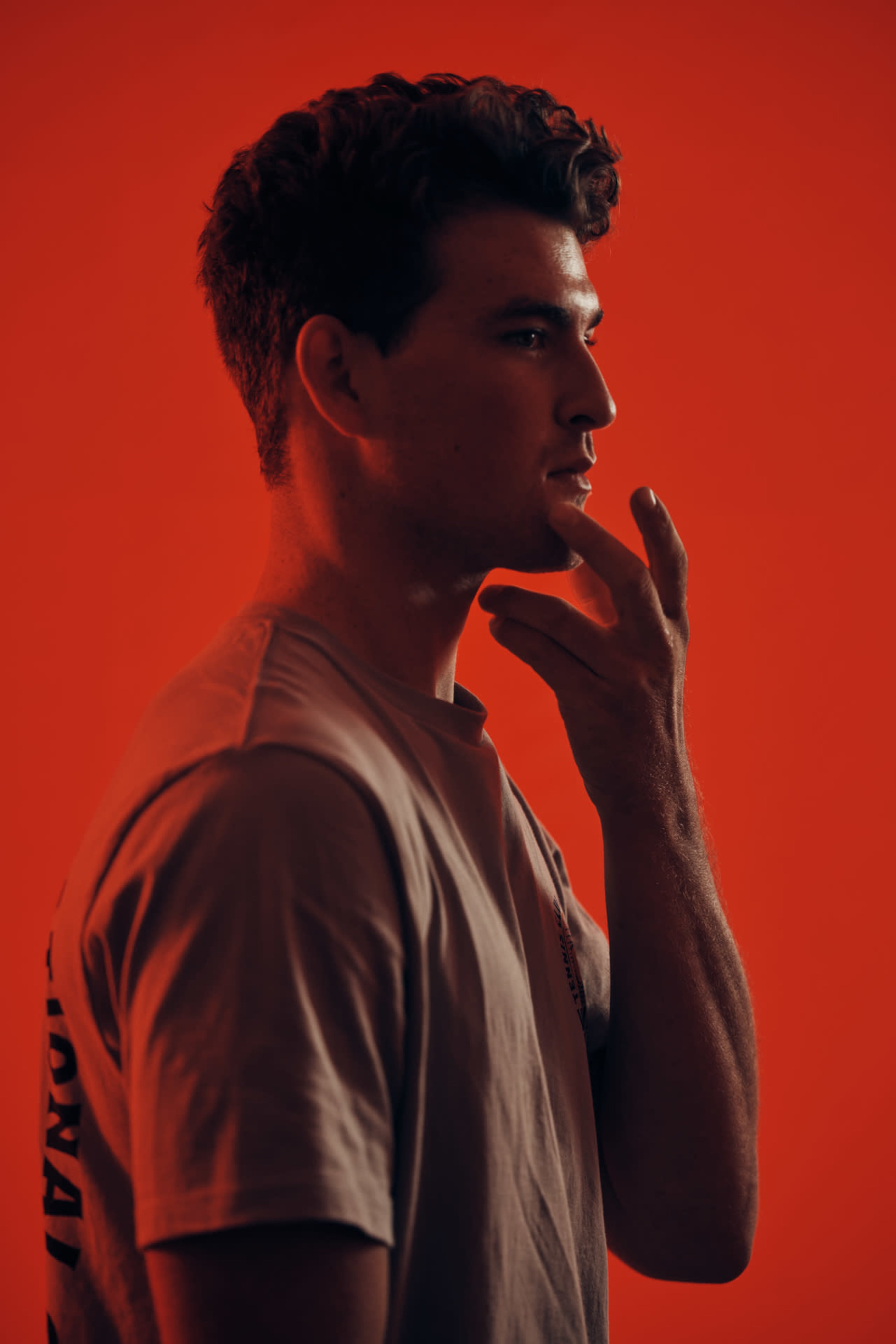 A man is standing in profile against a red background, touching his chin with one hand.