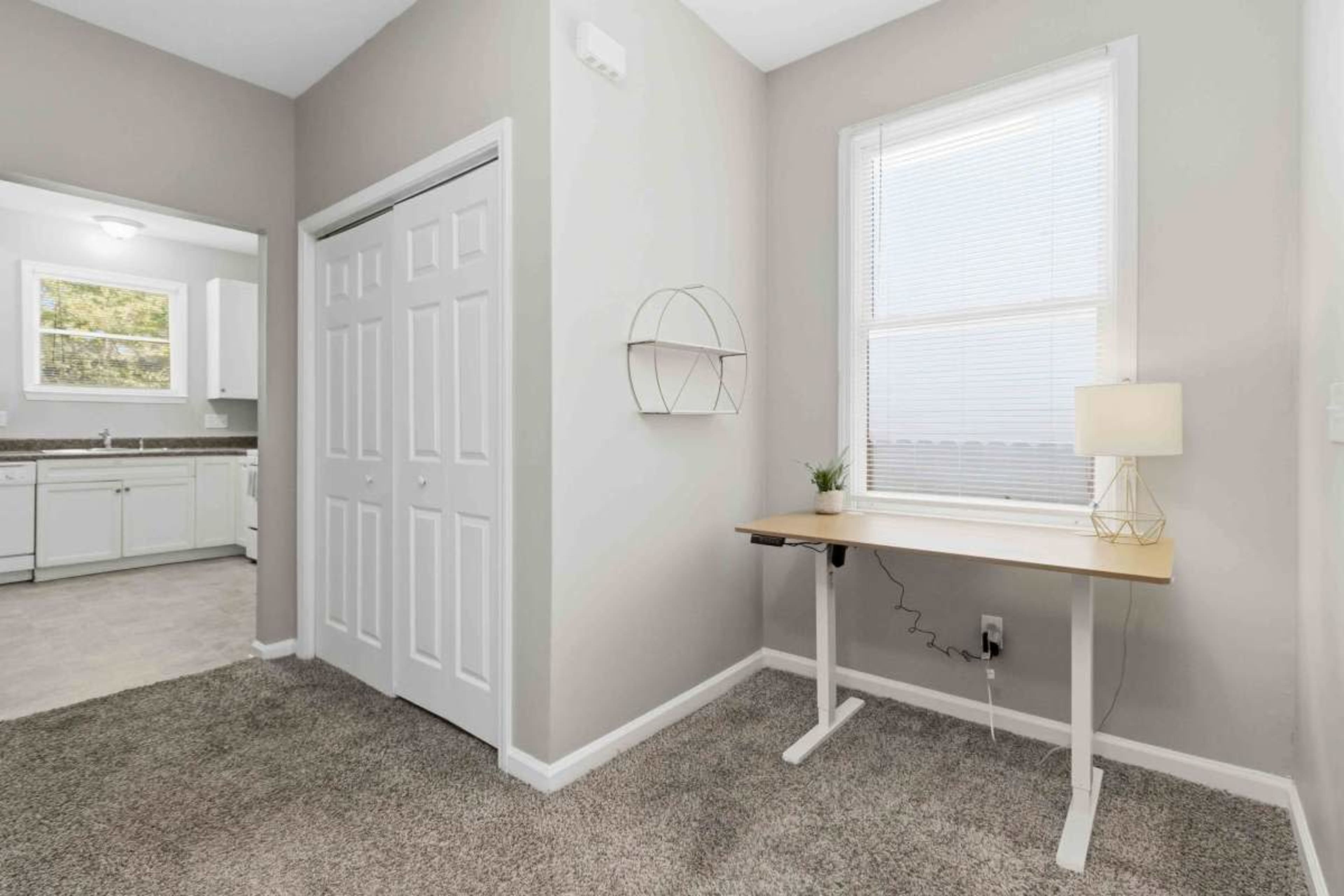 The image shows a small, well-lit room with a desk and lamp, leading into a kitchen area visible through a doorway.