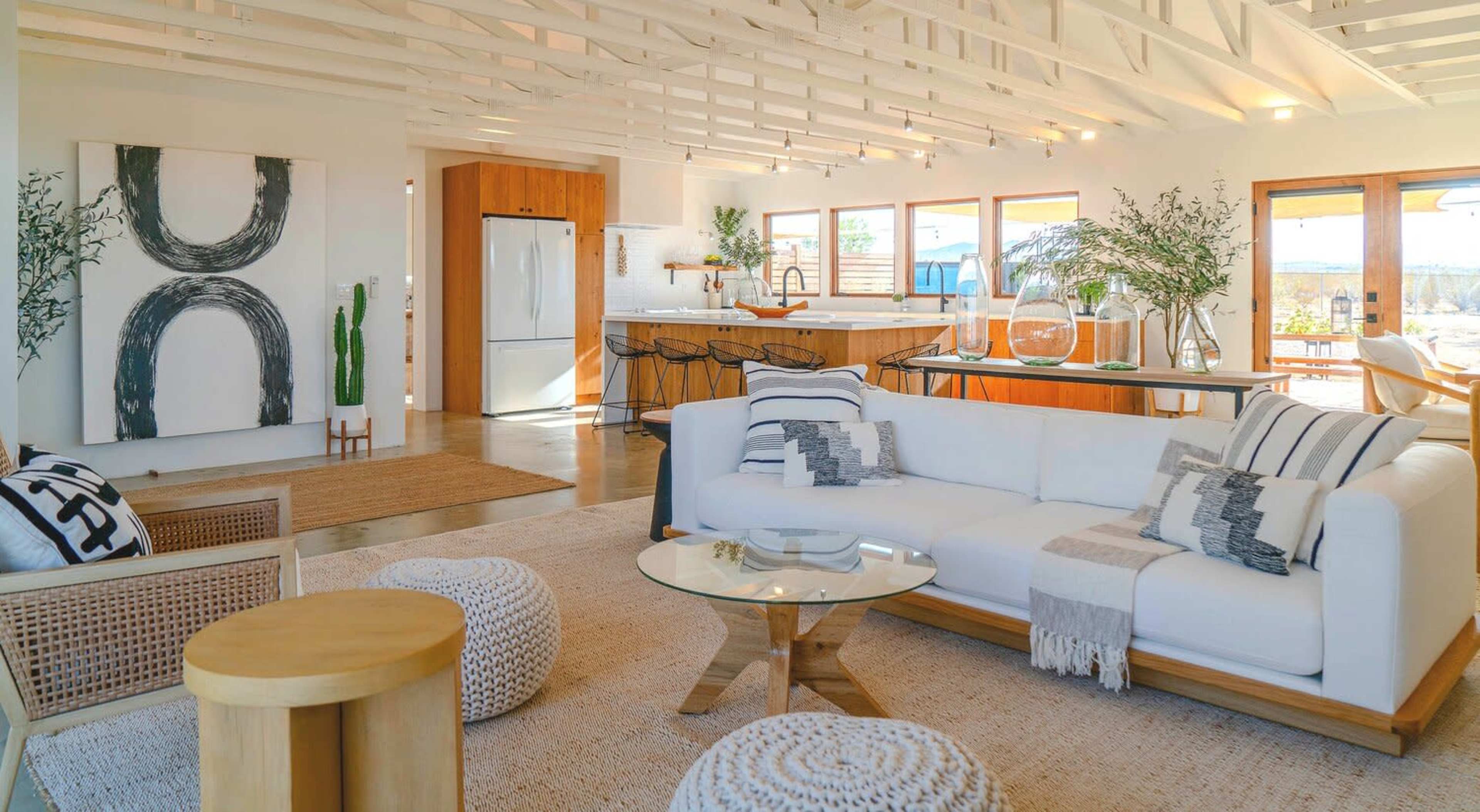 A modern living space features a white sofa, a glass coffee table, and a kitchen area with wooden accents and large windows.