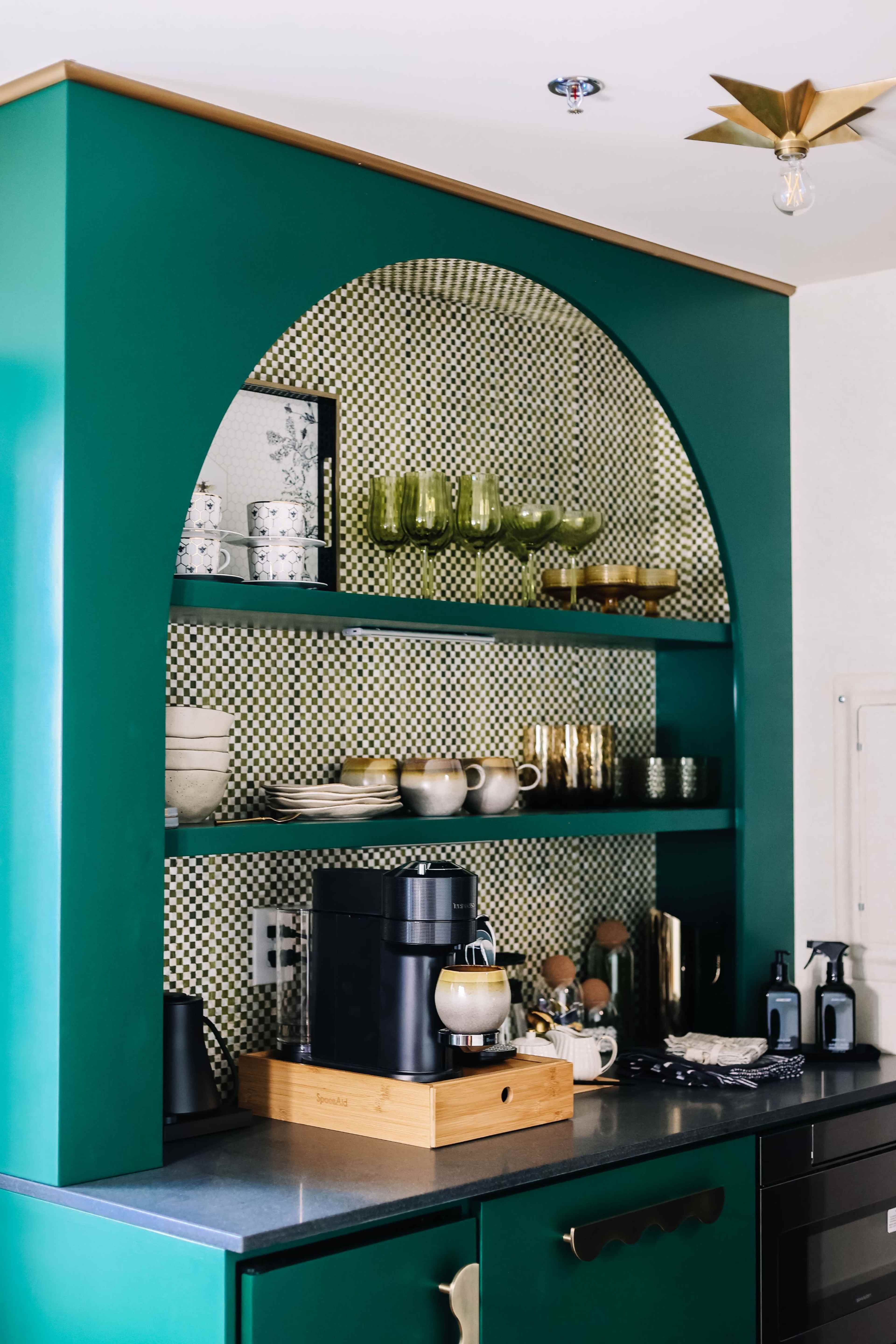The image shows a modern kitchen featuring a green archway cabinet with open shelves displaying various kitchenware and a coffee machine on a wooden stand.
