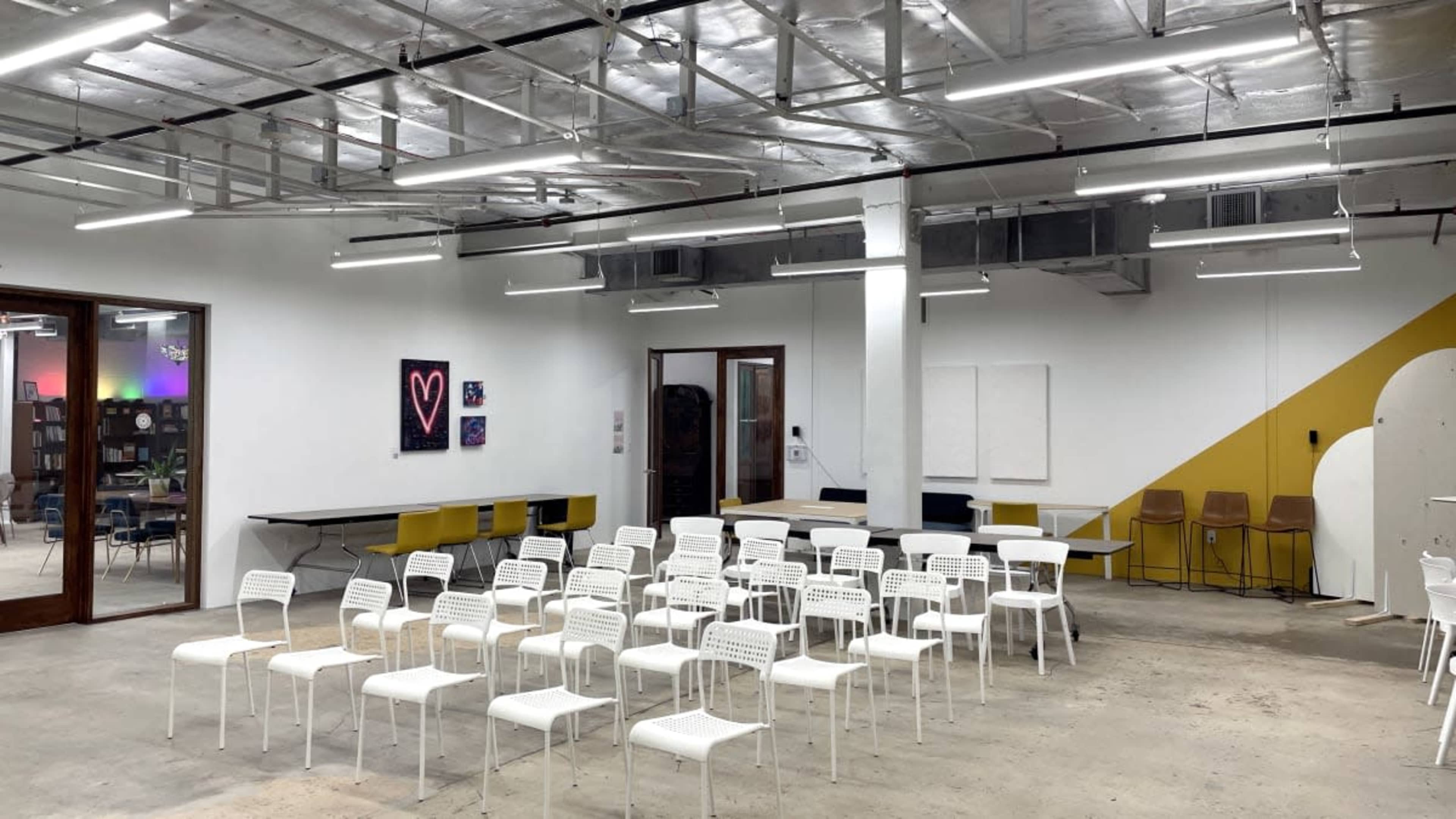 The image shows a spacious, well-lit room with rows of white chairs facing a white wall, alongside tables and a few colorful artworks.