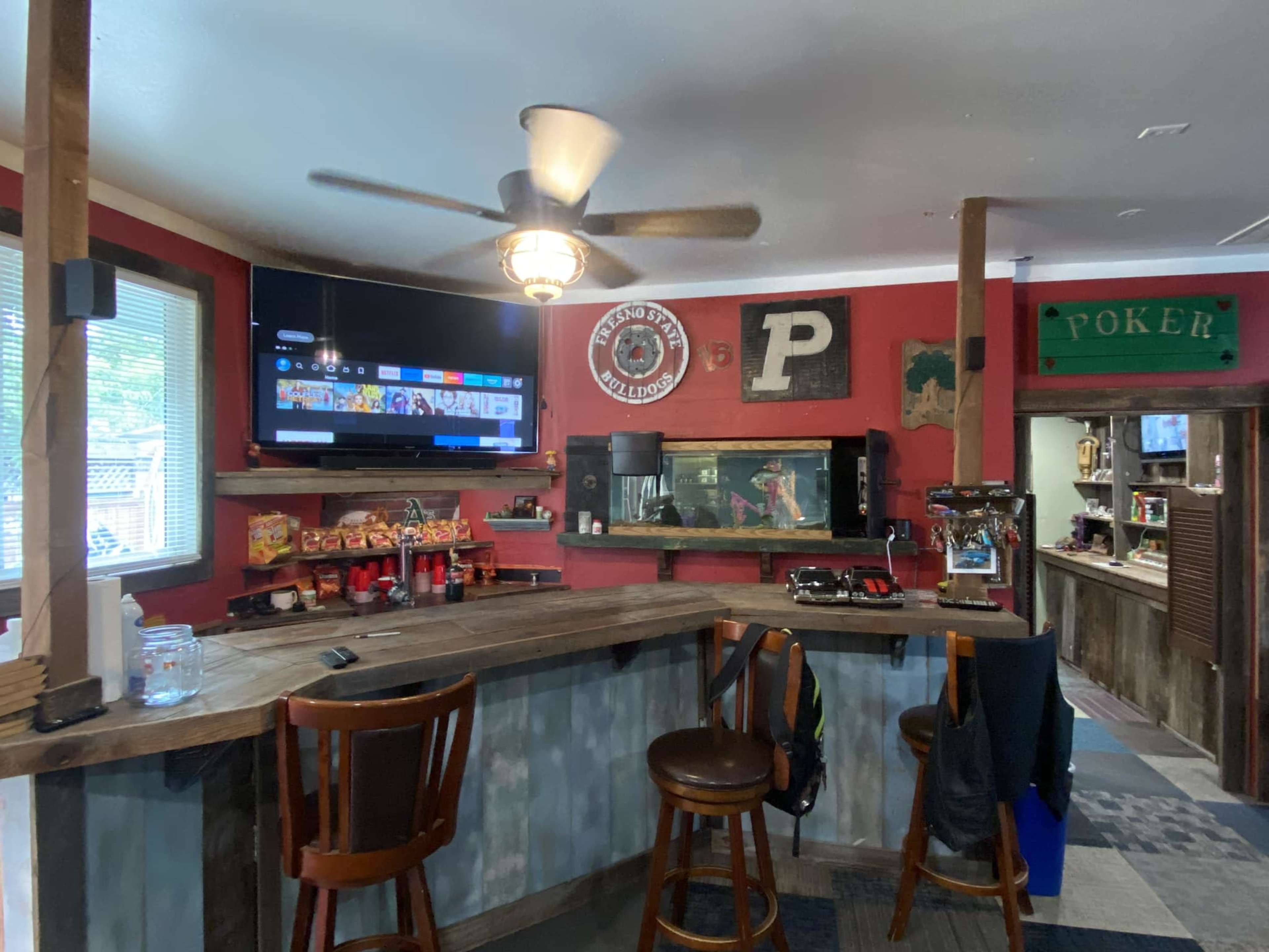 The image shows a cozy bar area with wooden stools, a TV mounted on the wall, and a variety of snacks displayed on the counter.
