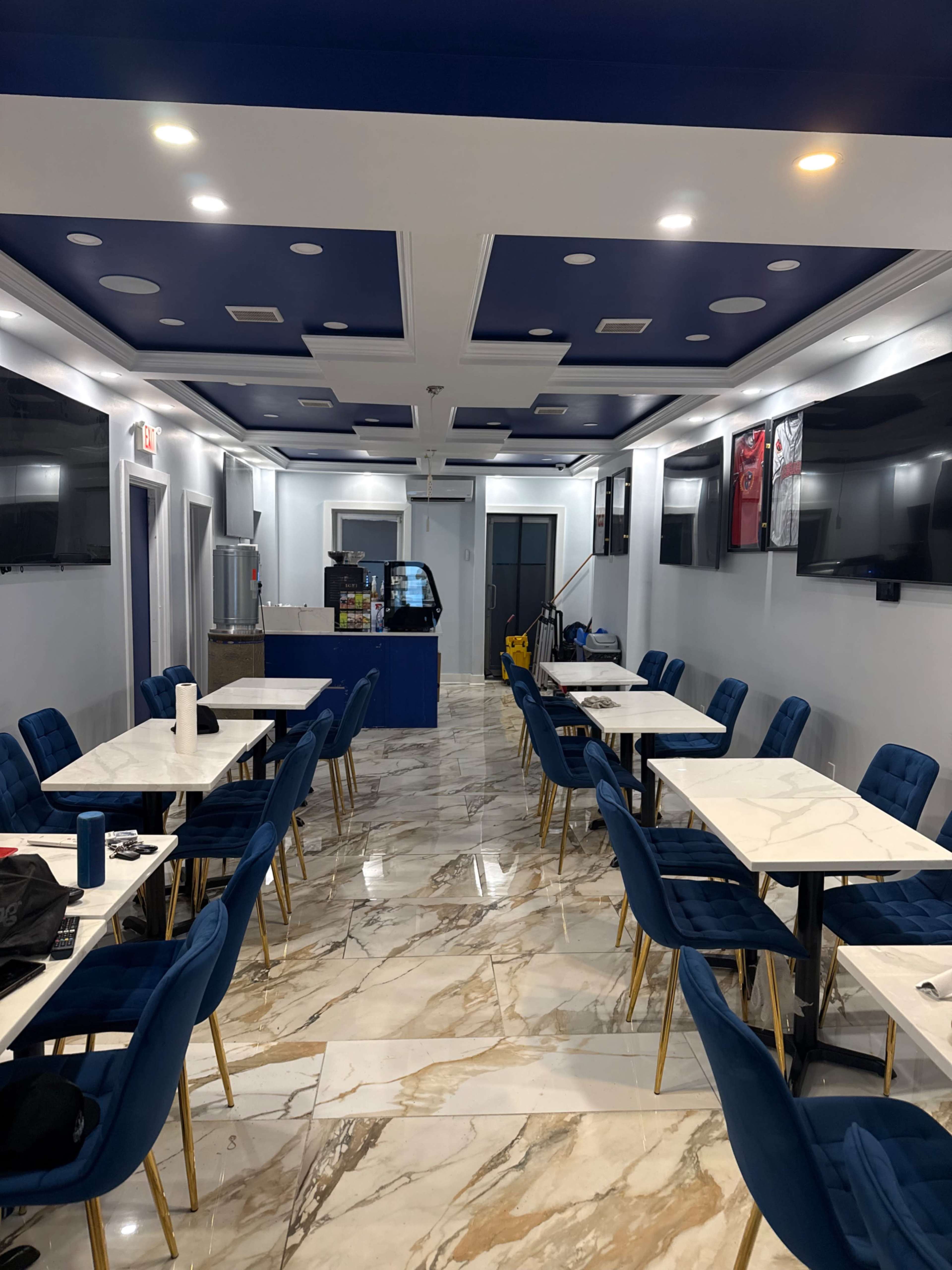 The image shows a modern café interior with blue velvet chairs and marble flooring, featuring tables arranged in a narrow layout and several wall-mounted televisions.