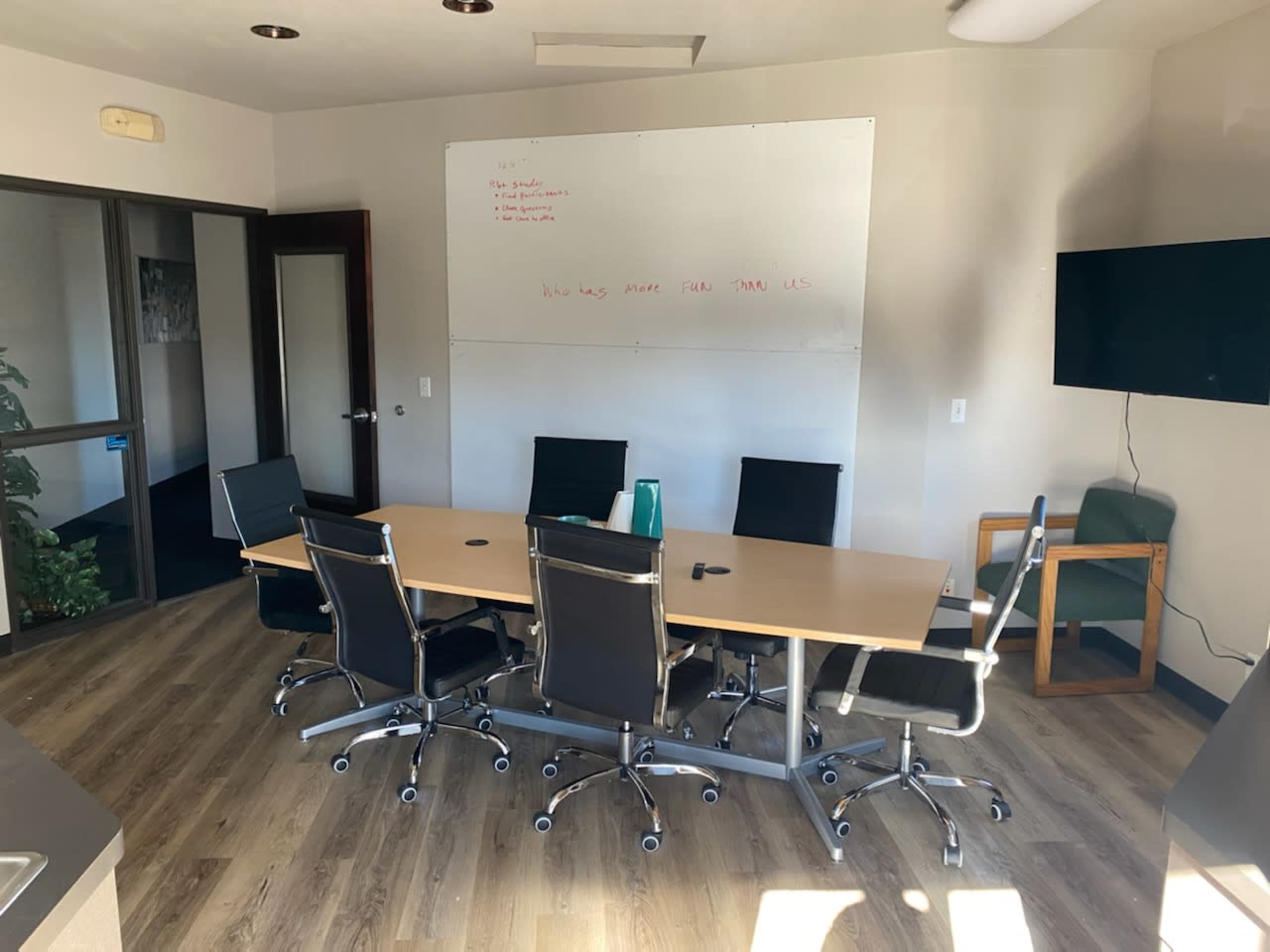A conference room features a wooden table surrounded by six black rolling chairs, a whiteboard on the wall, and a television mounted in one corner.
