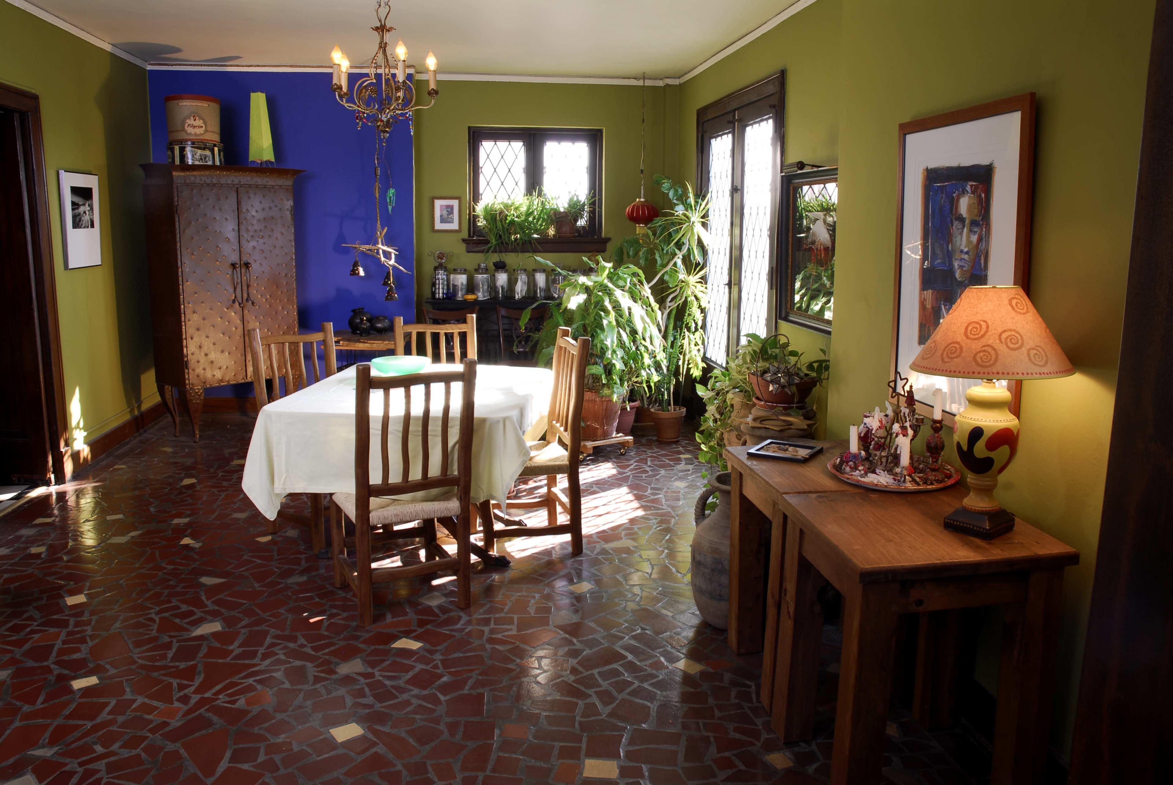 A dining area with a wooden table and chairs surrounded by green and blue walls, with plants and decorative items positioned throughout the room.