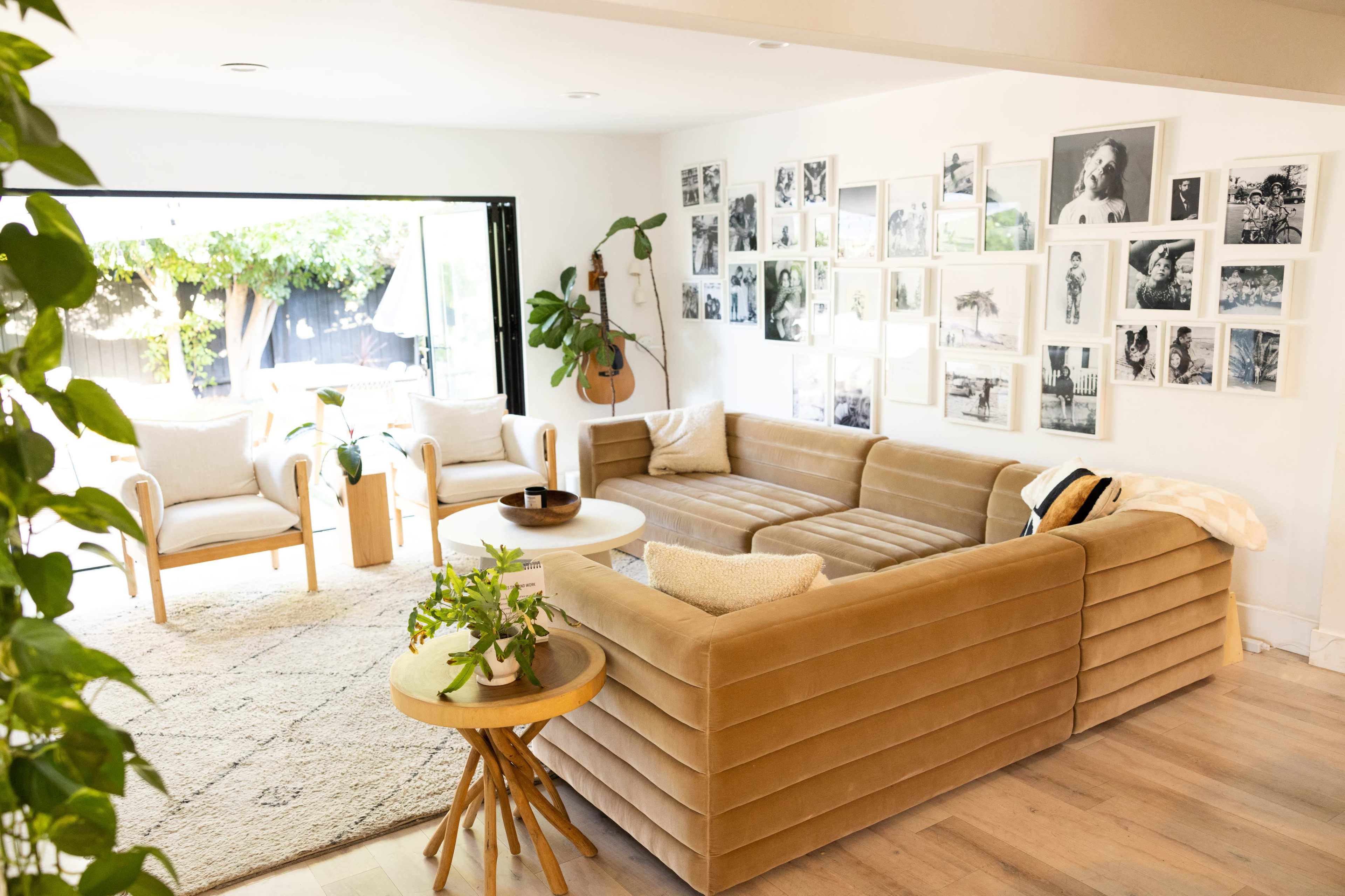The image shows a bright living room with a beige sectional couch, a round coffee table, and several framed black-and-white photographs hanging on the wall.