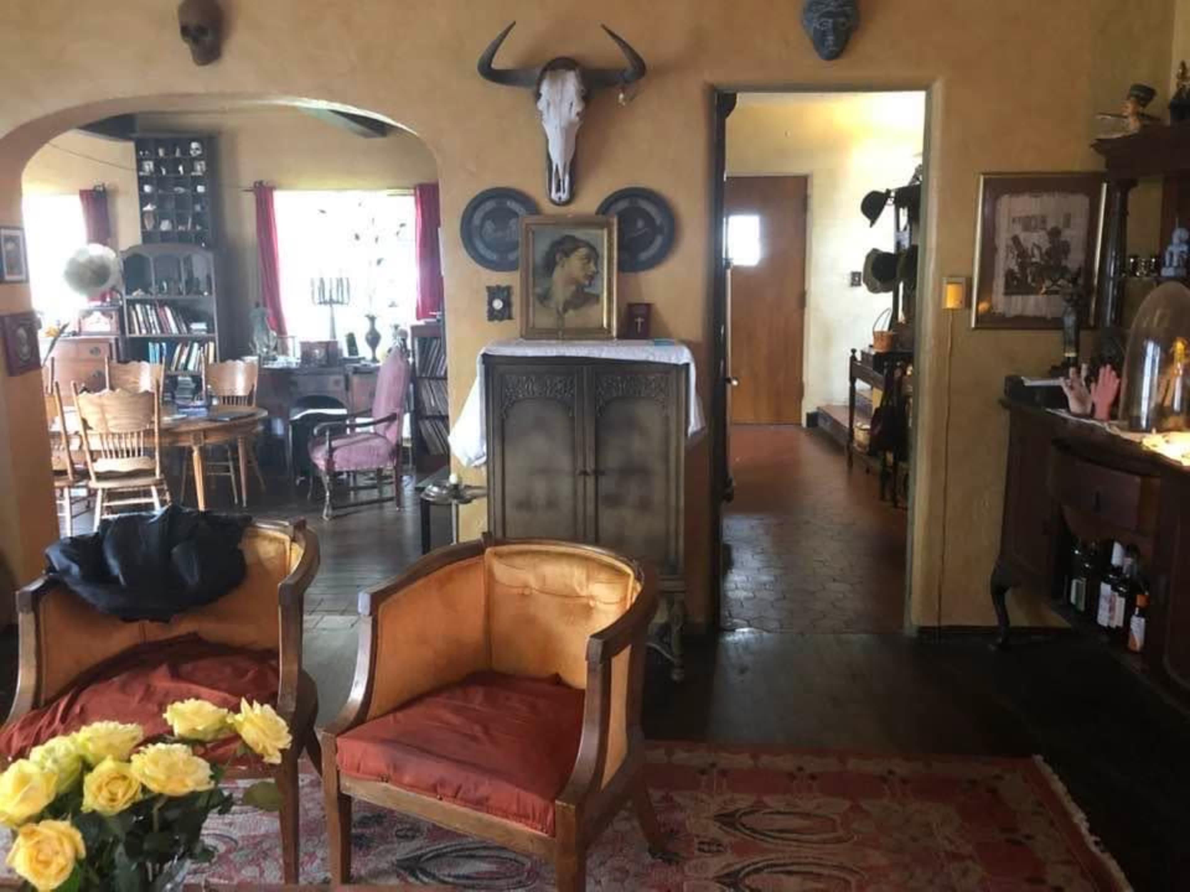 A warmly decorated room featuring two orange armchairs, a wooden dining table with chairs, and a hallway leading to another room, adorned with various wall decorations and a mounted animal skull.